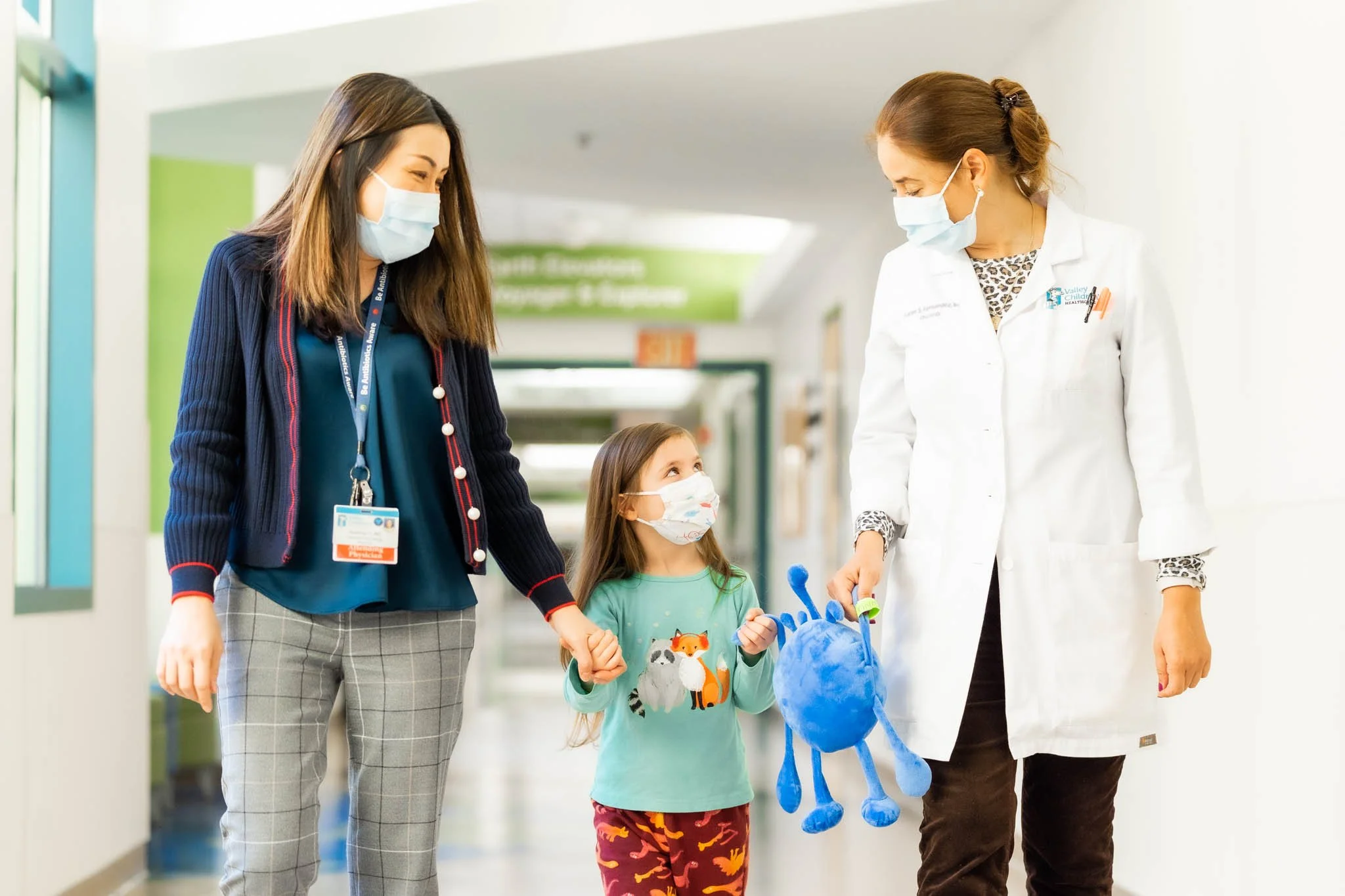 A young girl walking in a hospital corridor holding hands with a woman and a healthcare professional. All three are wearing face masks. The girl is carrying a blue stuffed toy.