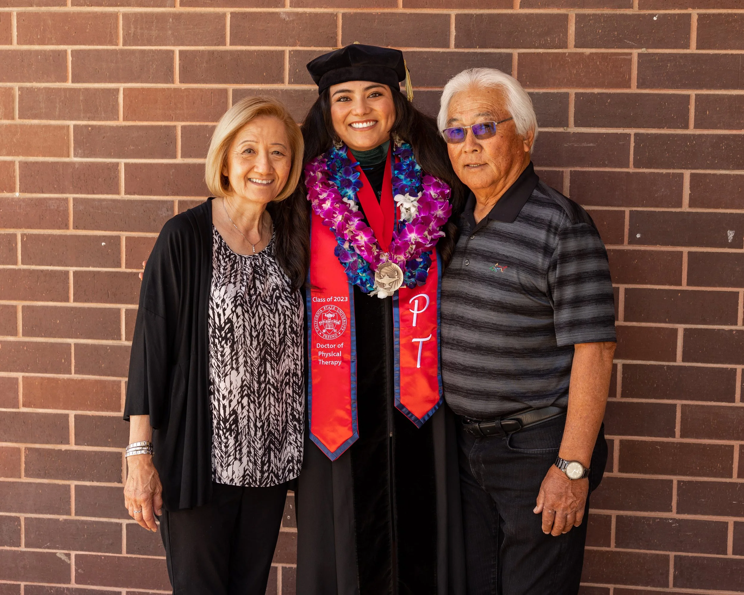 A young woman in graduation regalia, including a black cap and gown, standing with an older woman on her left and an older man on her right in front of a brick wall. The woman wears a red graduation stole with purple and blue leis and medals, indicat