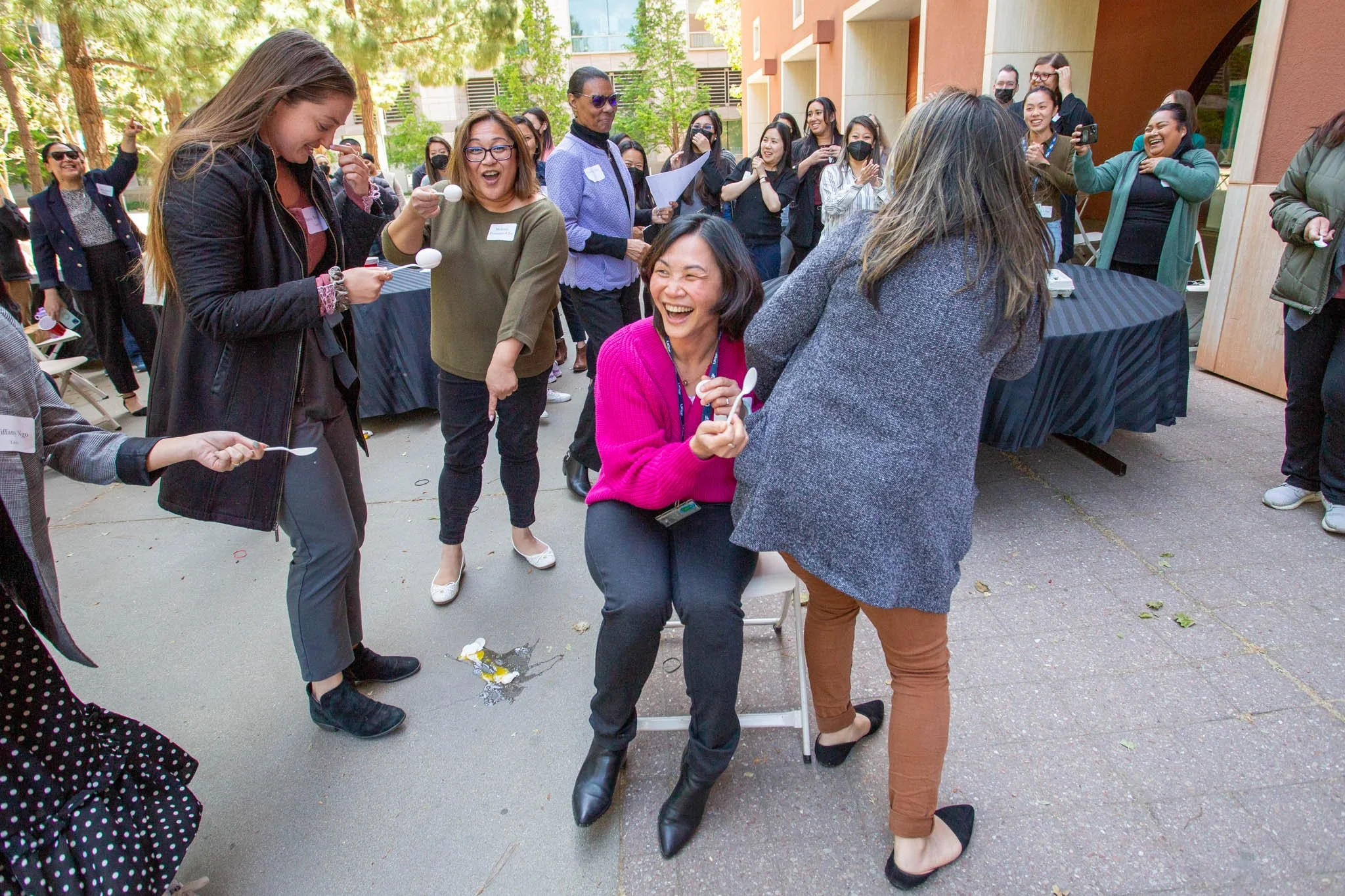 Group of women laughing and enjoying an outdoor event, some wearing masks, with a woman in a pink sweater sitting on a chair in the center.