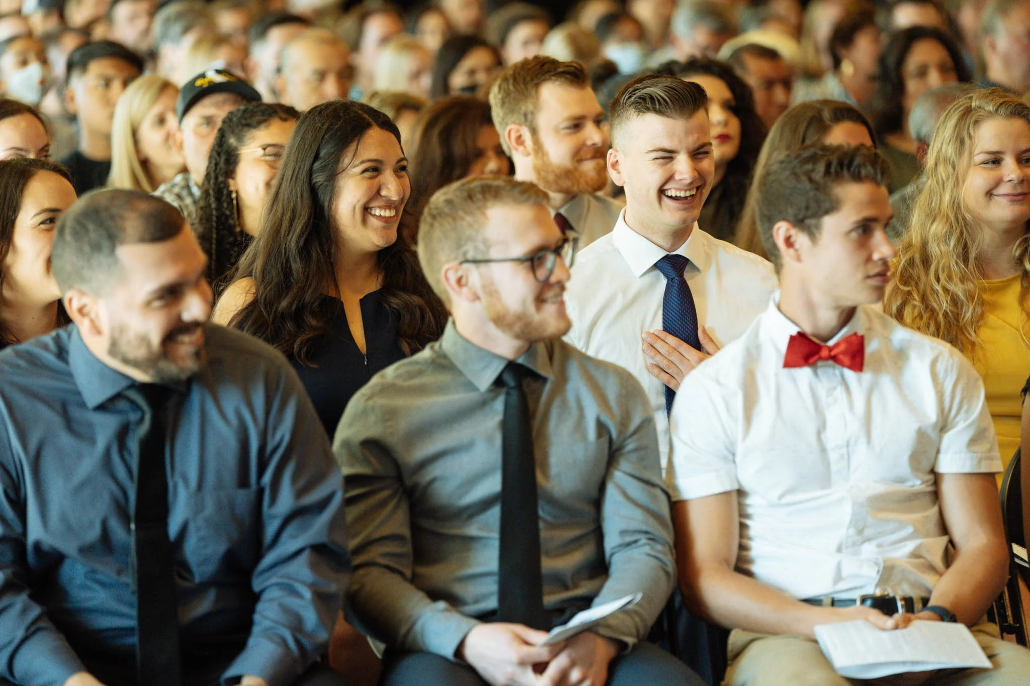 A group of diverse young adults attending an event, seated in rows, smiling and laughing, dressed in business casual and semi-formal attire.