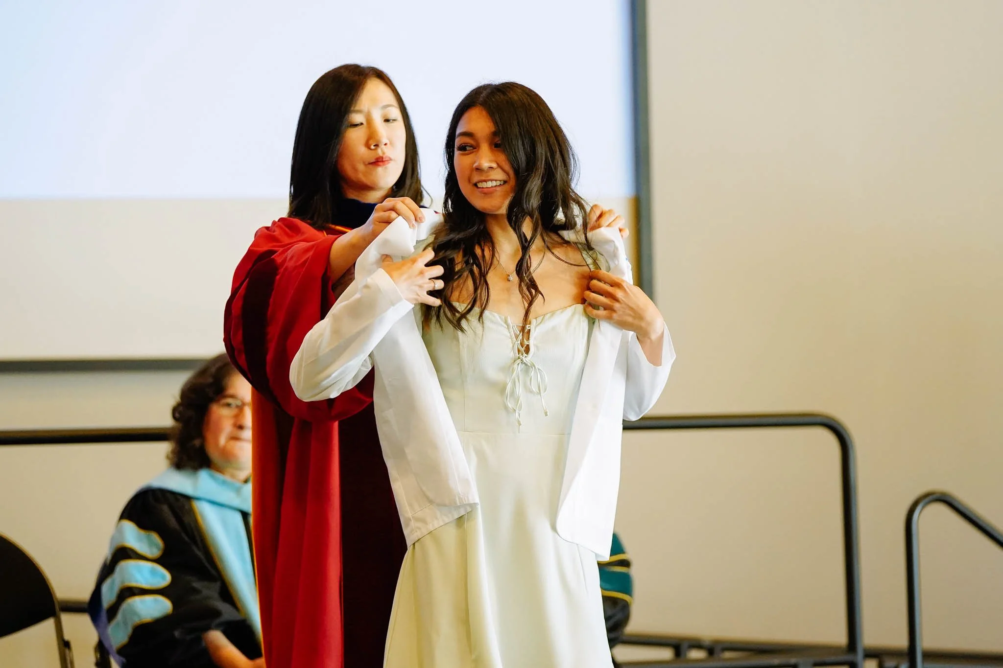 A graduate from california university of fresno is being assisted by another woman in her graduation gown at a ceremony. An audience member watches in the background.