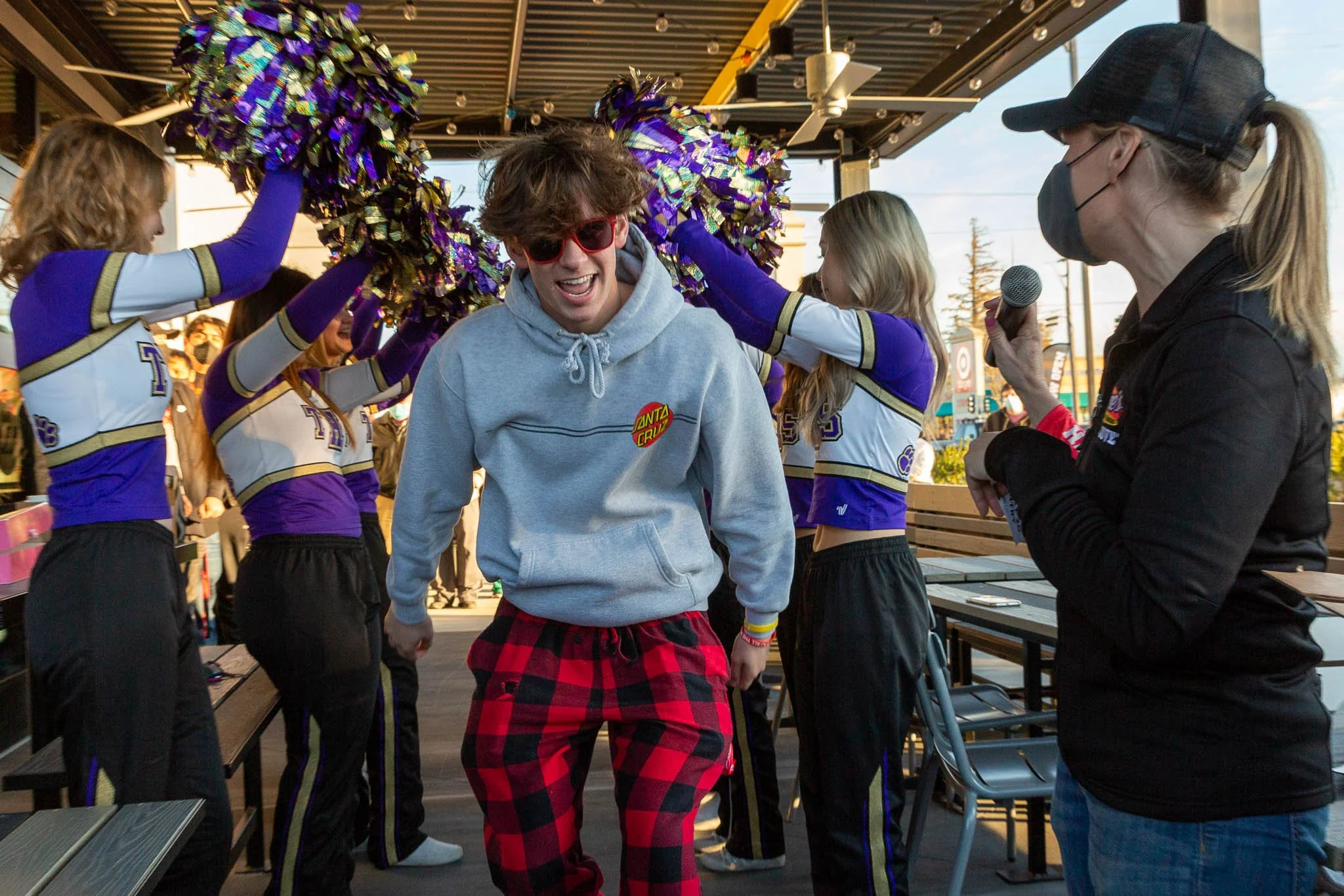 A young man with curly hair, sunglasses, a gray hoodie, and red plaid pants is smiling while dancing with cheerleaders holding pom-poms at an outdoor event, with a woman wearing a face mask and holding a microphone talking to him.