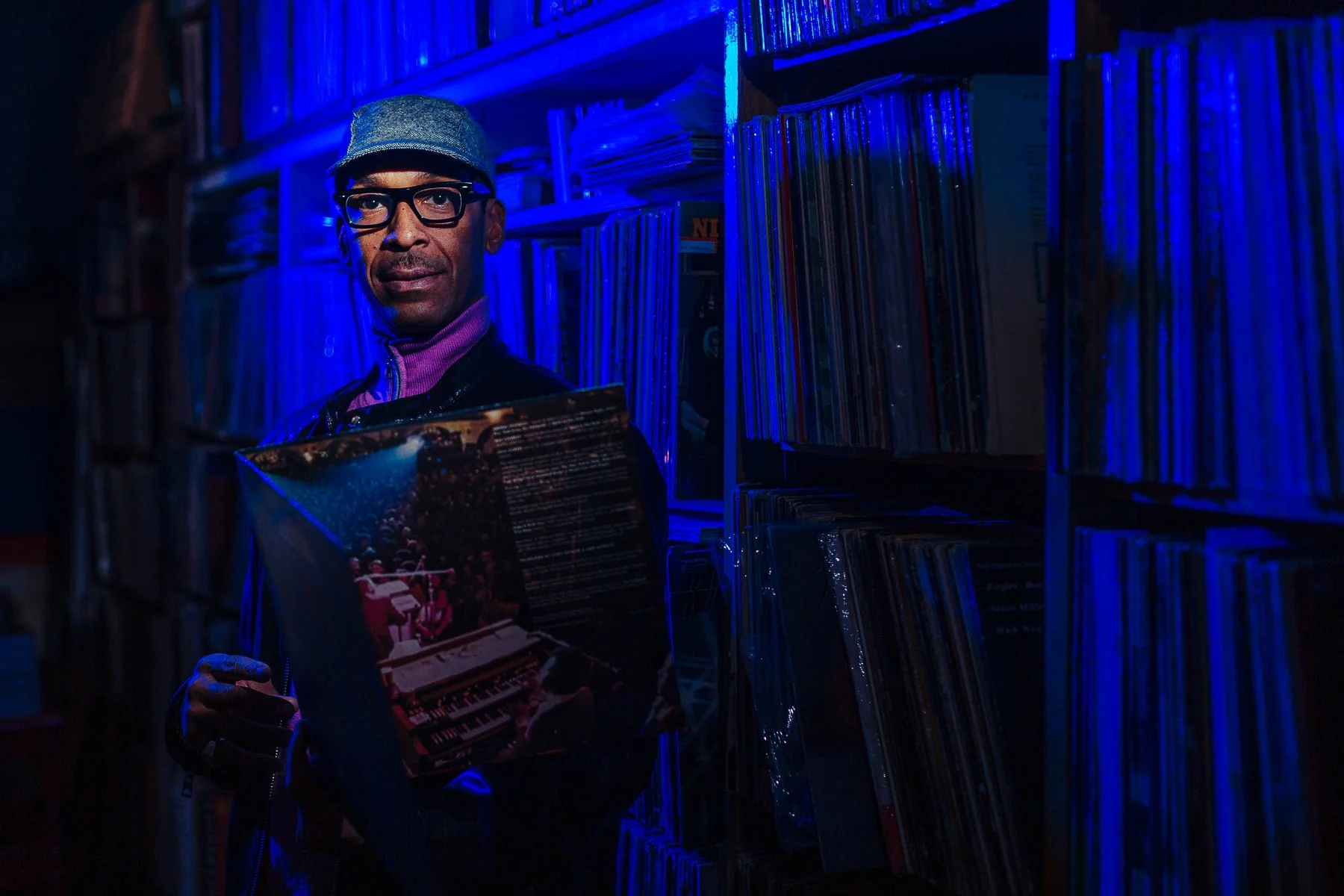 A man wearing glasses, a cap, and a purple turtleneck stands in a dimly lit record store, holding a record album with a colorful cover. The store has shelves filled with vinyl records, illuminated by blue lighting.