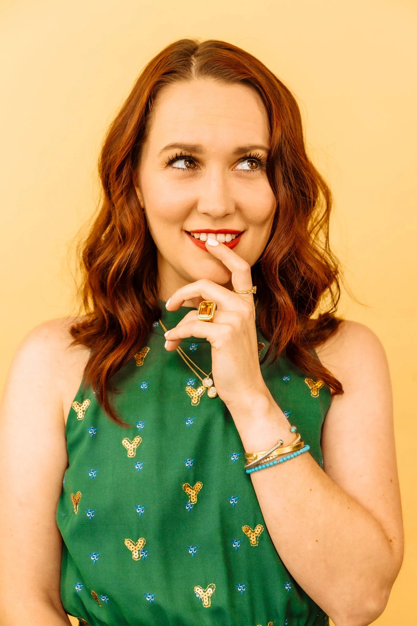 A woman with red hair, wearing a green sleeveless dress with butterfly and floral print, smiling and holding her finger to her lips, standing against a yellow background.