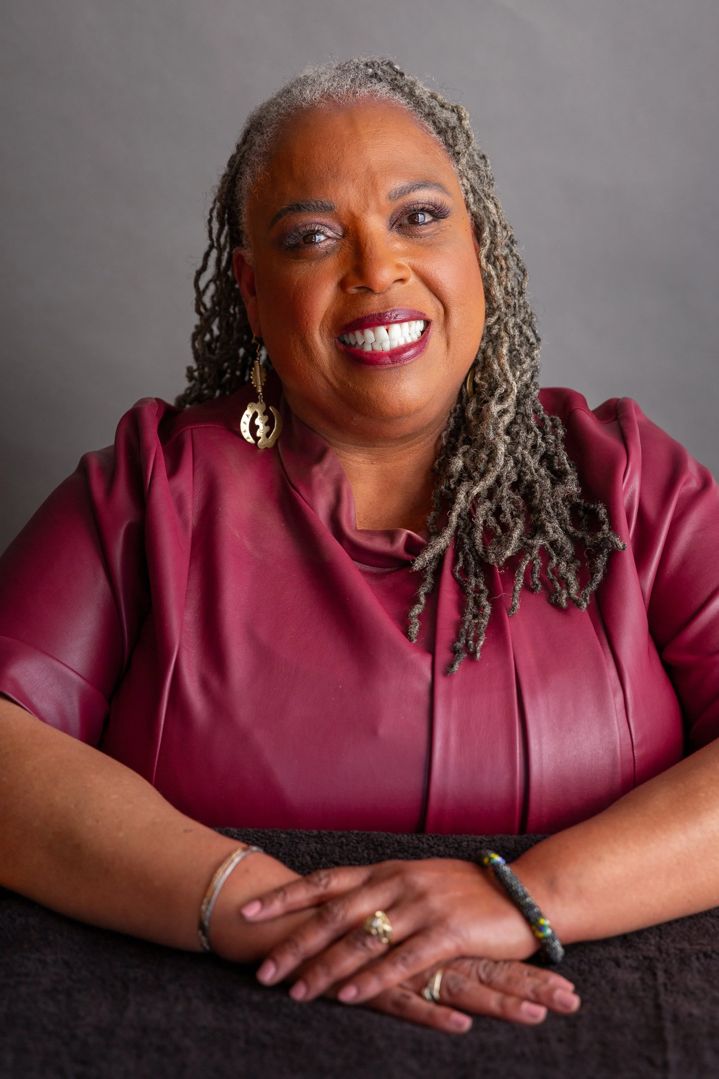 A smiling African American woman with gray dreadlocks wearing a burgundy leather top and gold earrings, sitting with her hands clasped on a dark surface, against a gray background.