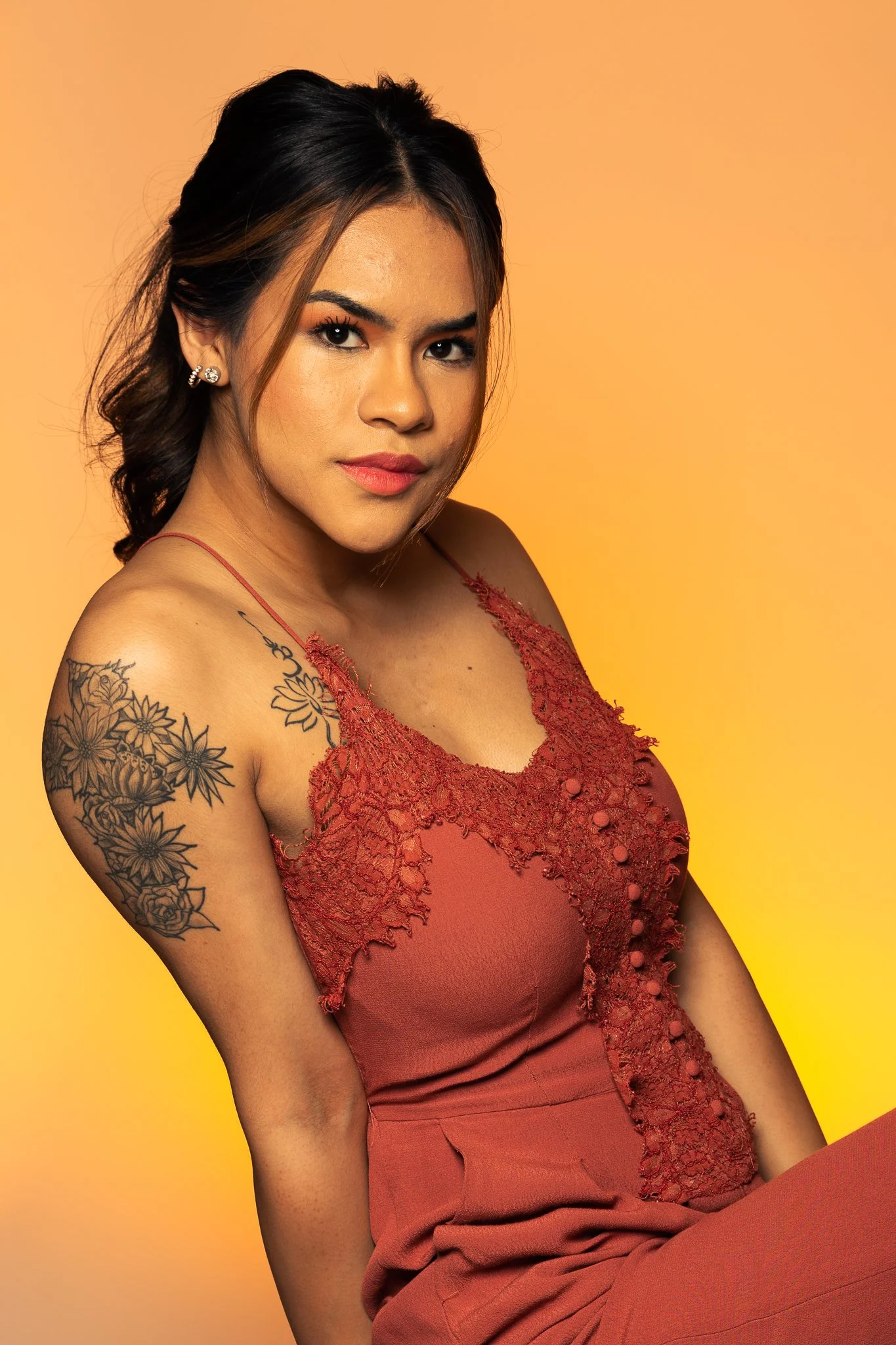 A young woman with dark hair styled in loose waves, wearing a spaghetti strap red dress with lace details, and earrings, posing against a yellow-orange background.