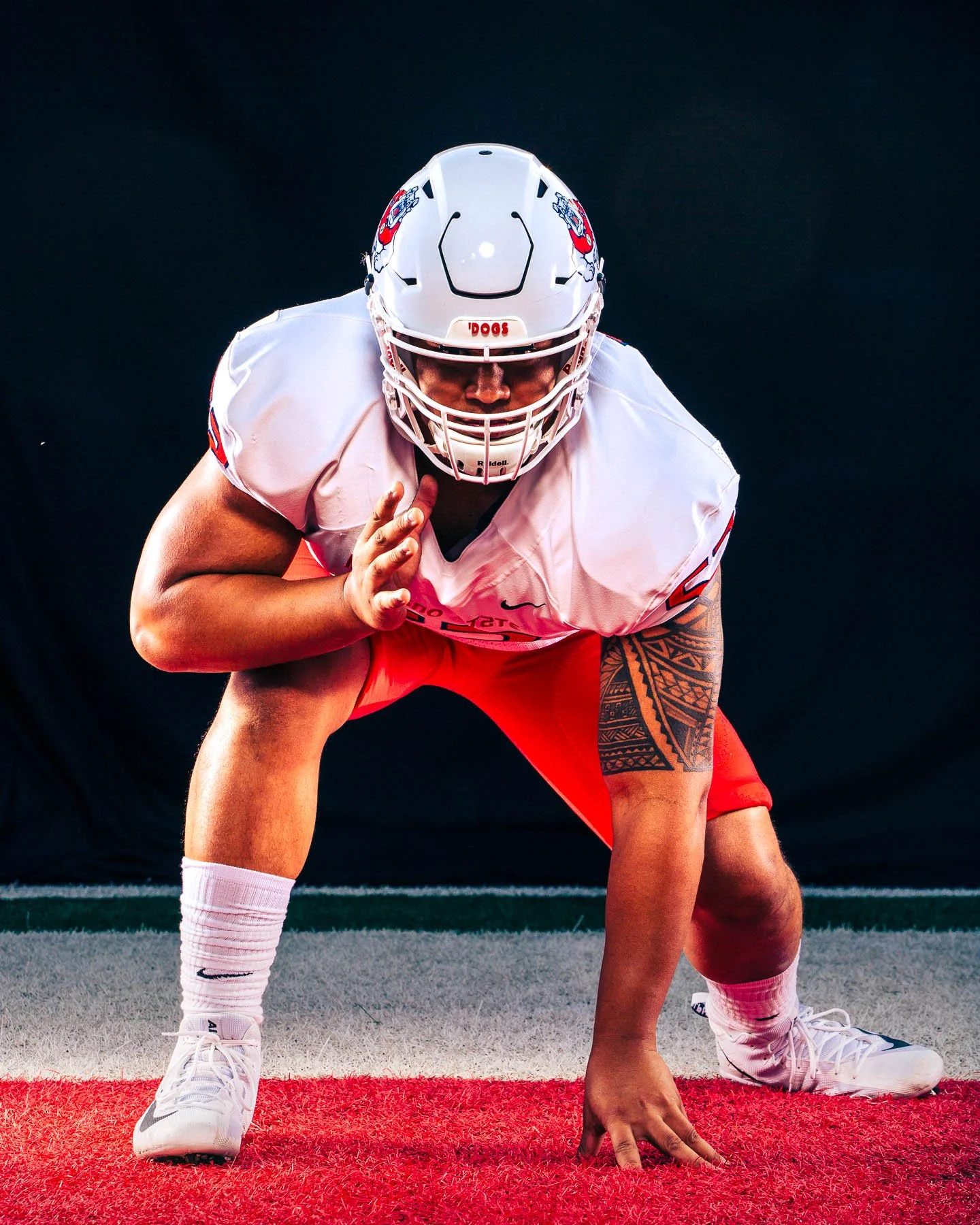 A football player in a white helmet and jersey crouches in a ready position on the field, with one hand on the ground and the other near his face mask. The background is dark, and the player is on a red and green field.