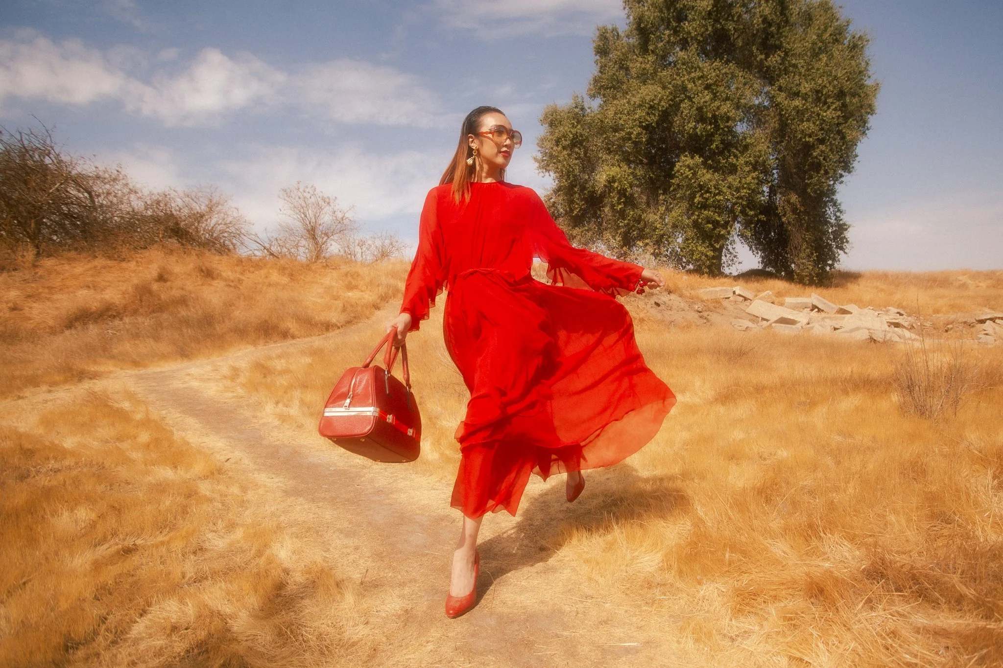 A woman in a flowing red dress and high heels is walking on a dirt path through a dry, grassy field, carrying a red handbag. She is wearing sunglasses and earrings, with a large tree and some rocks in the background under a partly cloudy sky.