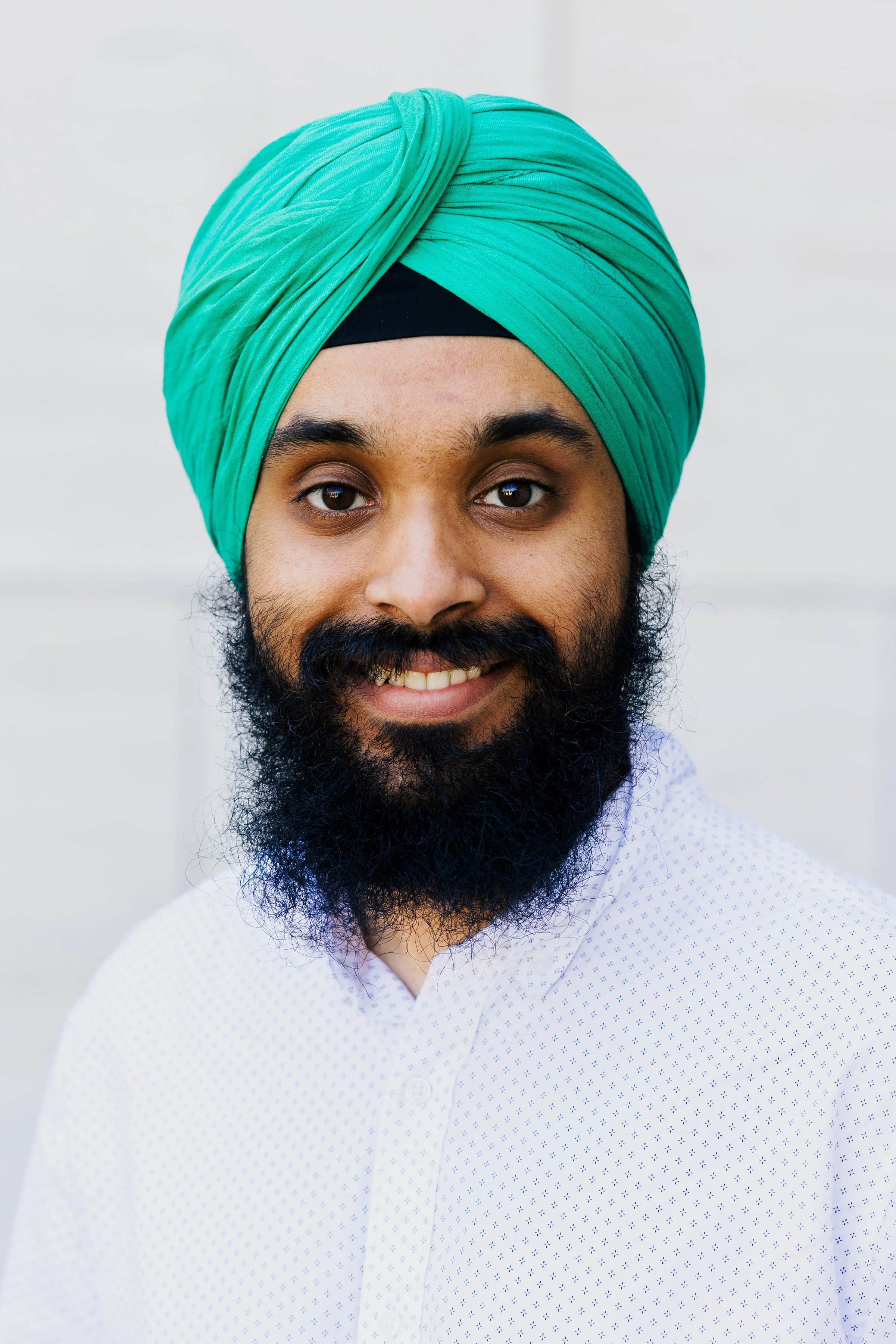 A man with a black beard and mustache, wearing a bright green turban and a white button-up shirt, smiling at the camera against a plain light background.