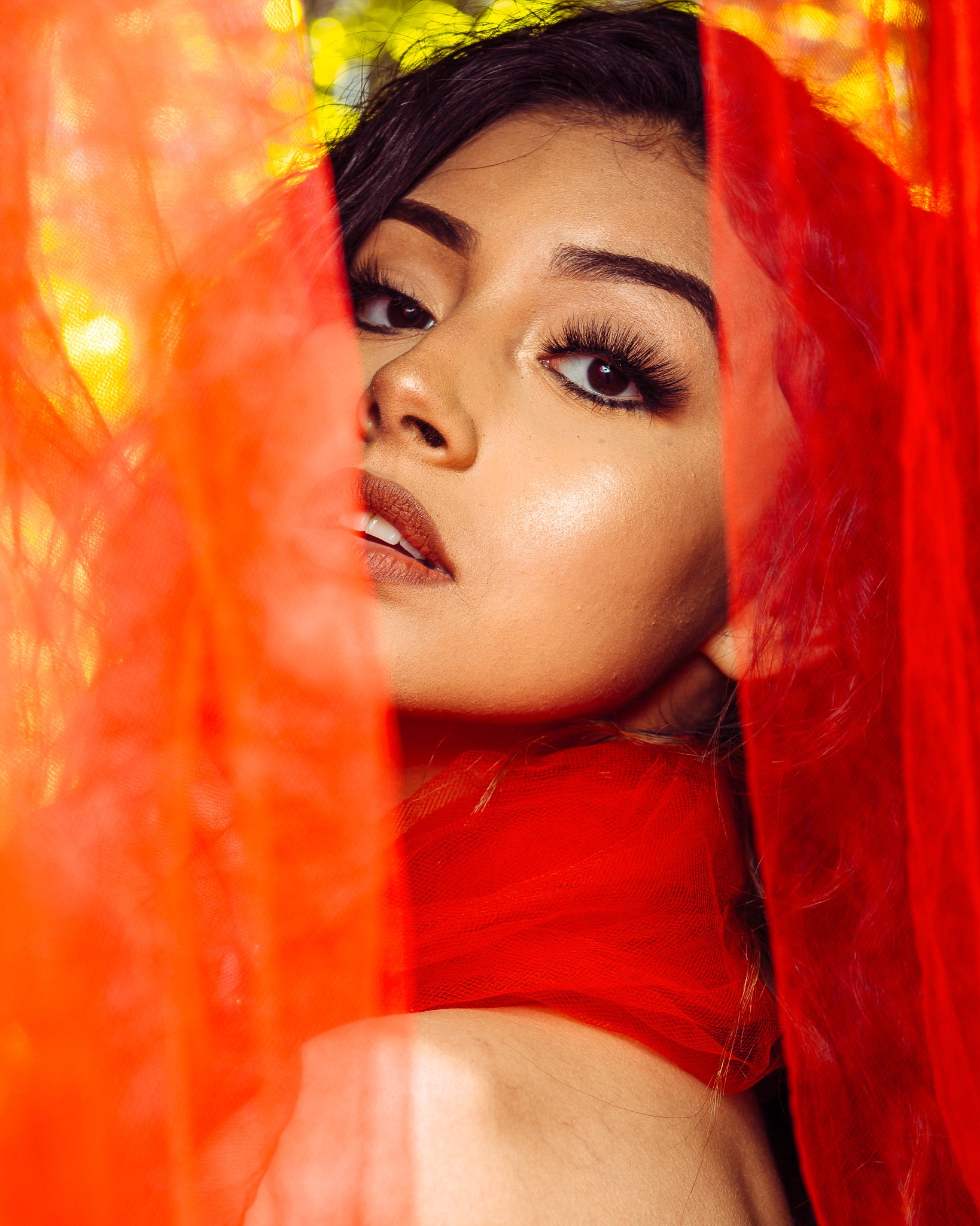 Close-up of a woman with dark hair and makeup, partially covered by red sheer fabric, with a blurred background of yellow and orange hues.
