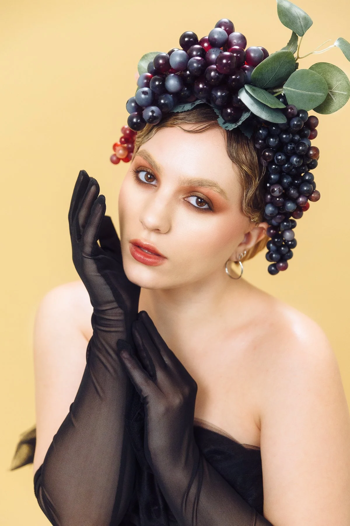 A woman with short curly hair and gold hoop earrings, wearing black sheer gloves, poses with her hand near her face against a yellow background. She is adorned with a crown made of grapes and green leaves on her head.