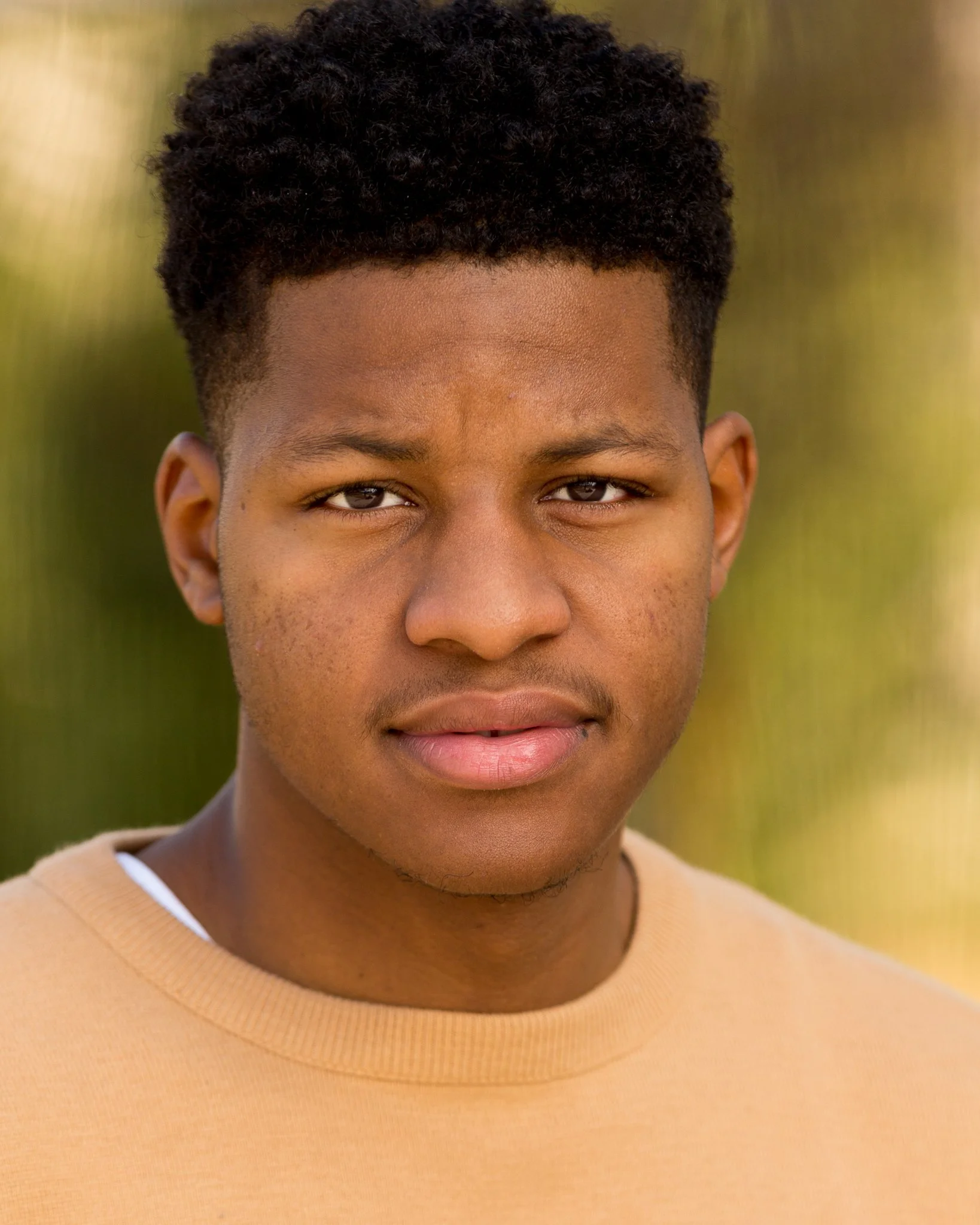 Close-up portrait of a young African American man with short curly hair, wearing a beige shirt, outdoors with a blurred green background.