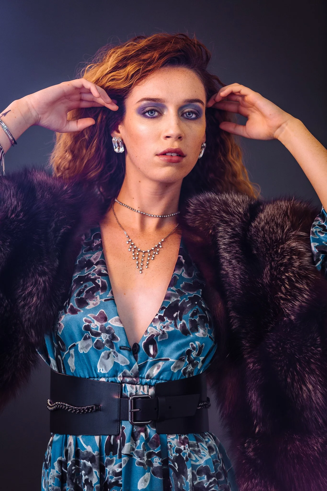 A woman with red wavy hair posing with her hands near her head, wearing a patterned dress, a wide black belt, a faux fur coat, and jewelry including earrings, a necklace, and bracelets.