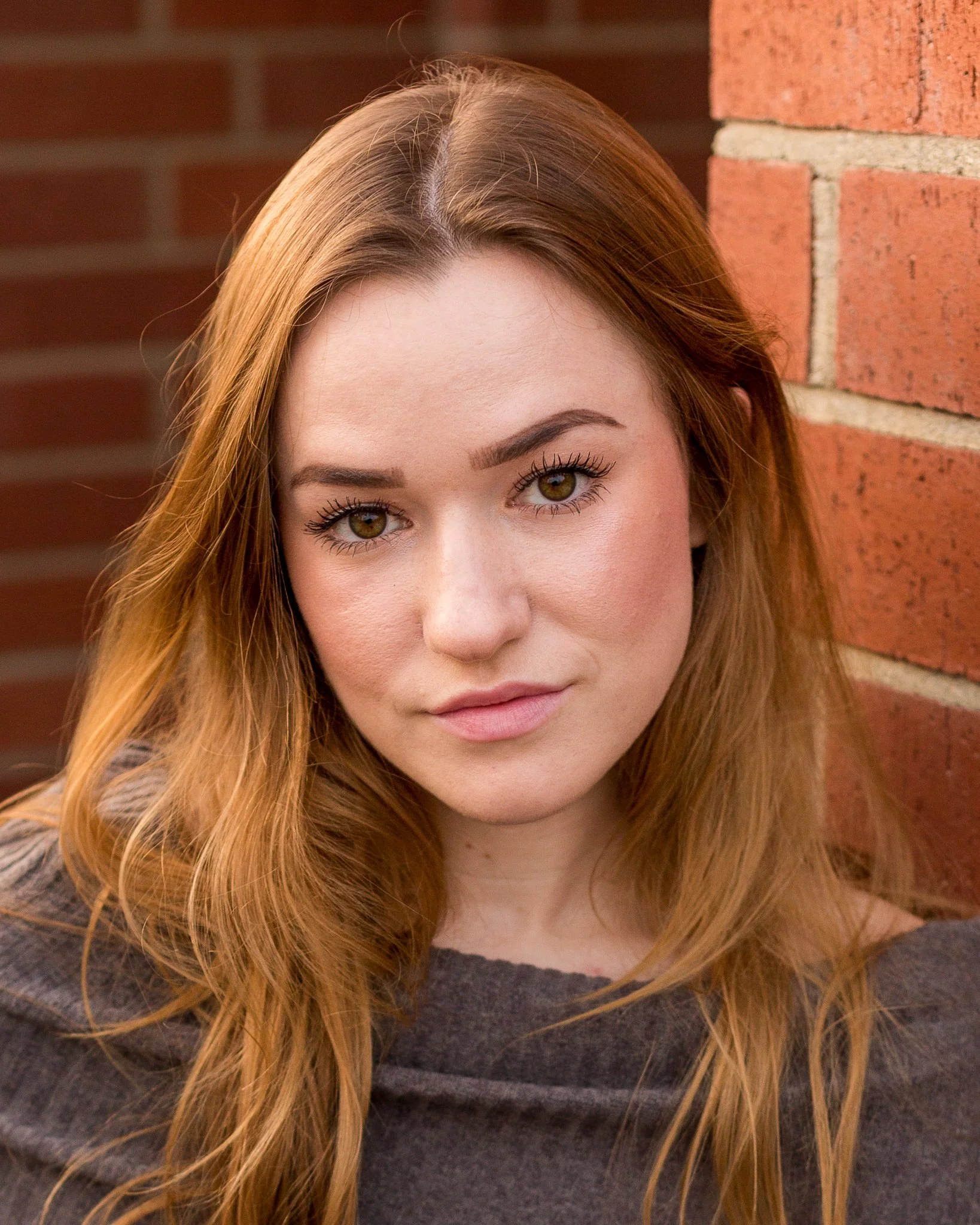 Close-up portrait of a young woman with red hair and light makeup, looking at the camera, with a brick wall background.