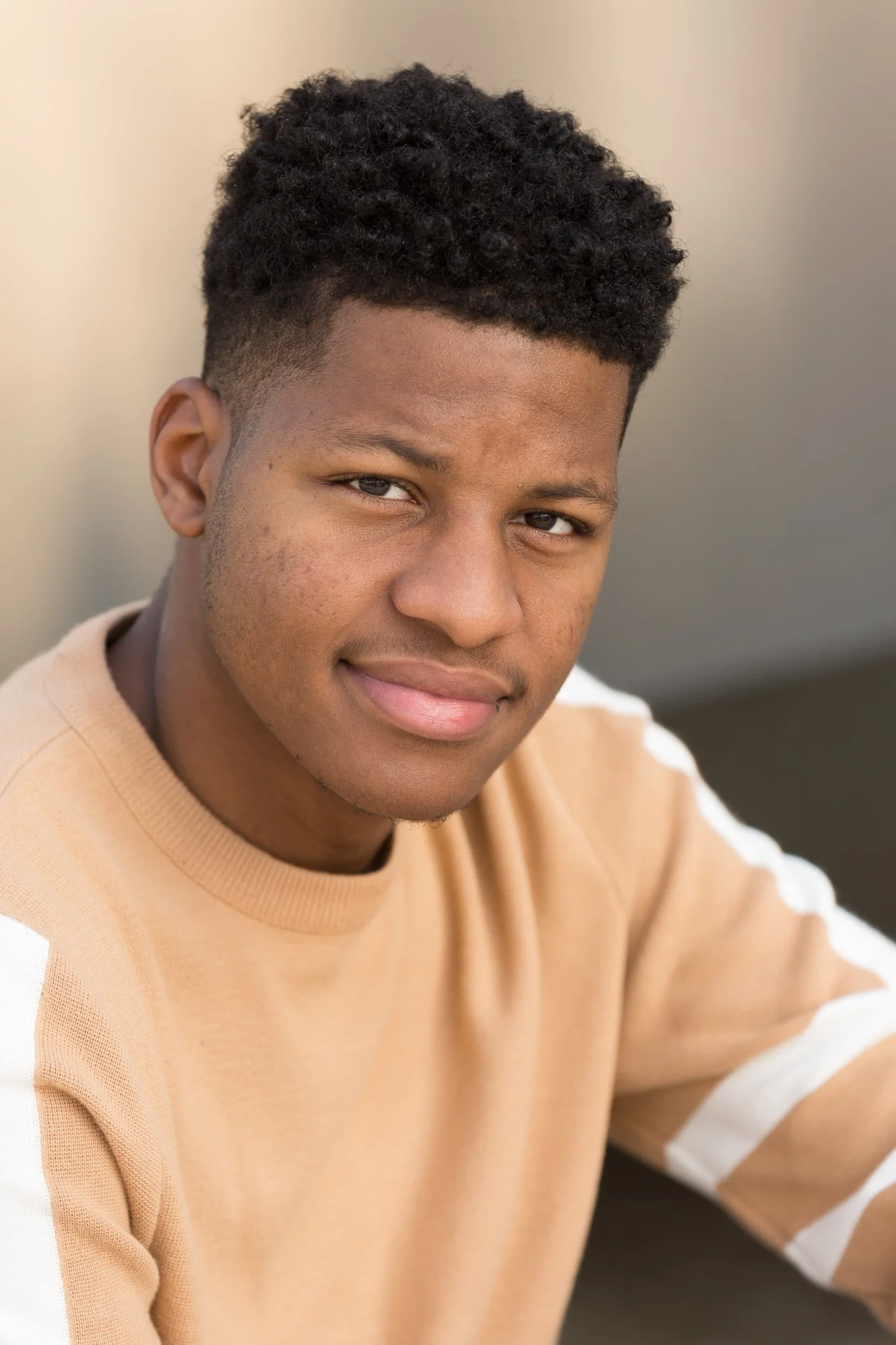 Close-up portrait of a young man with short curly hair, wearing a beige sweatshirt with white stripes on the sleeves, smiling subtly.