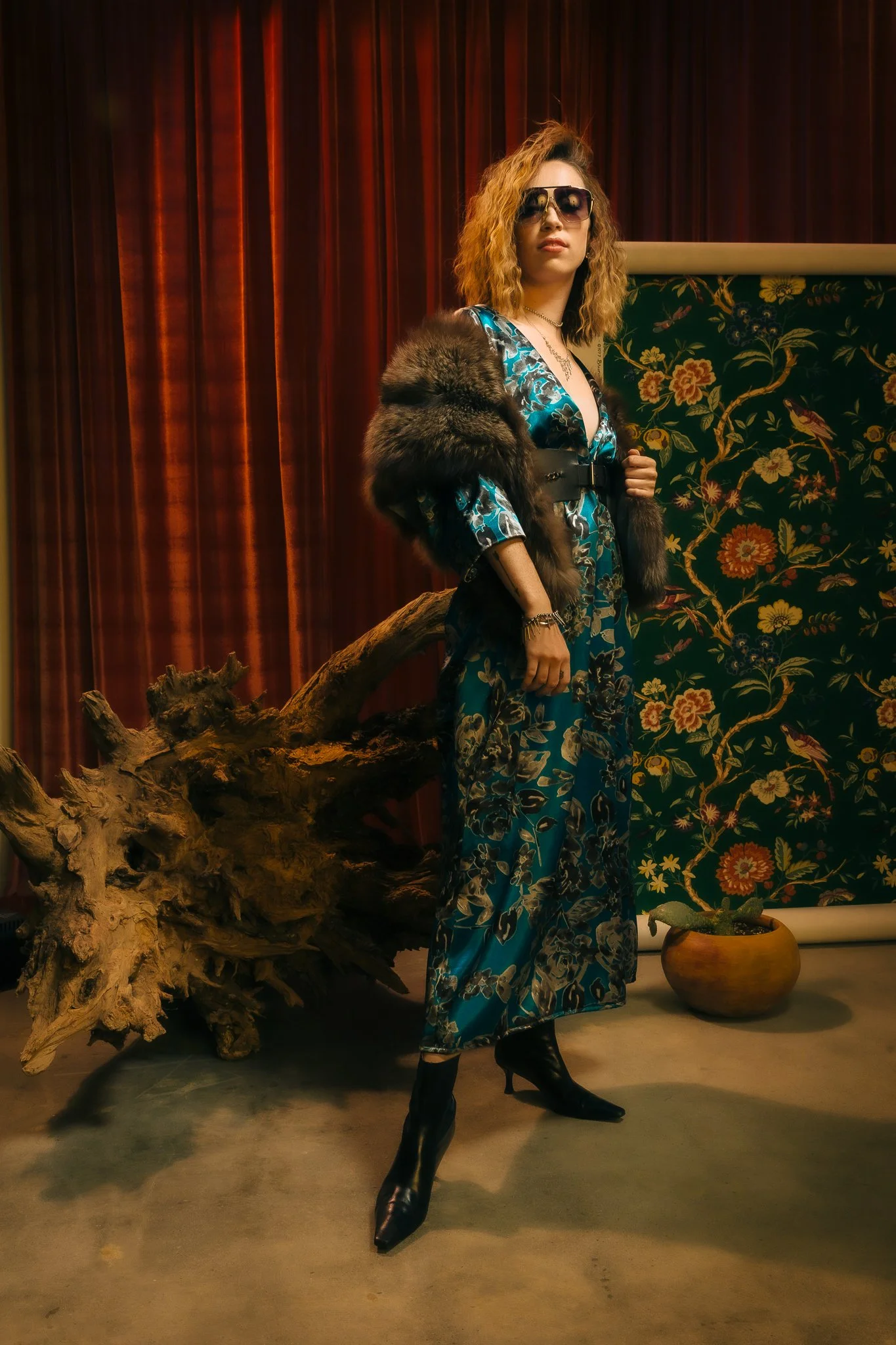A woman with curly hair wearing sunglasses, a floral jumpsuit, a fur stole, and black heeled boots, standing in front of a decorative background with red curtains, a floral tapestry, a large piece of driftwood, and a potted plant.
