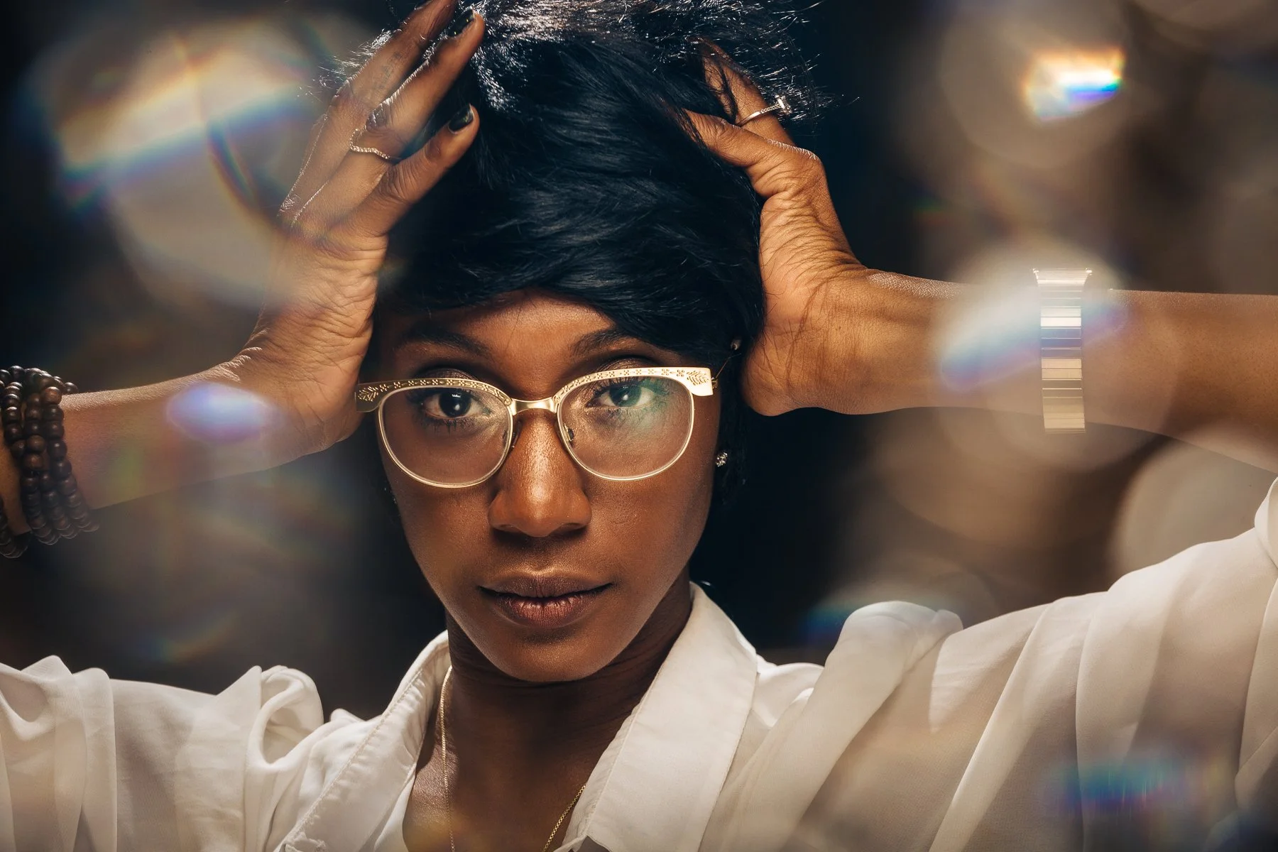 A woman with short black hair, glasses, and a white blouse, holding her head with her hands amidst a hazy, colorful background.