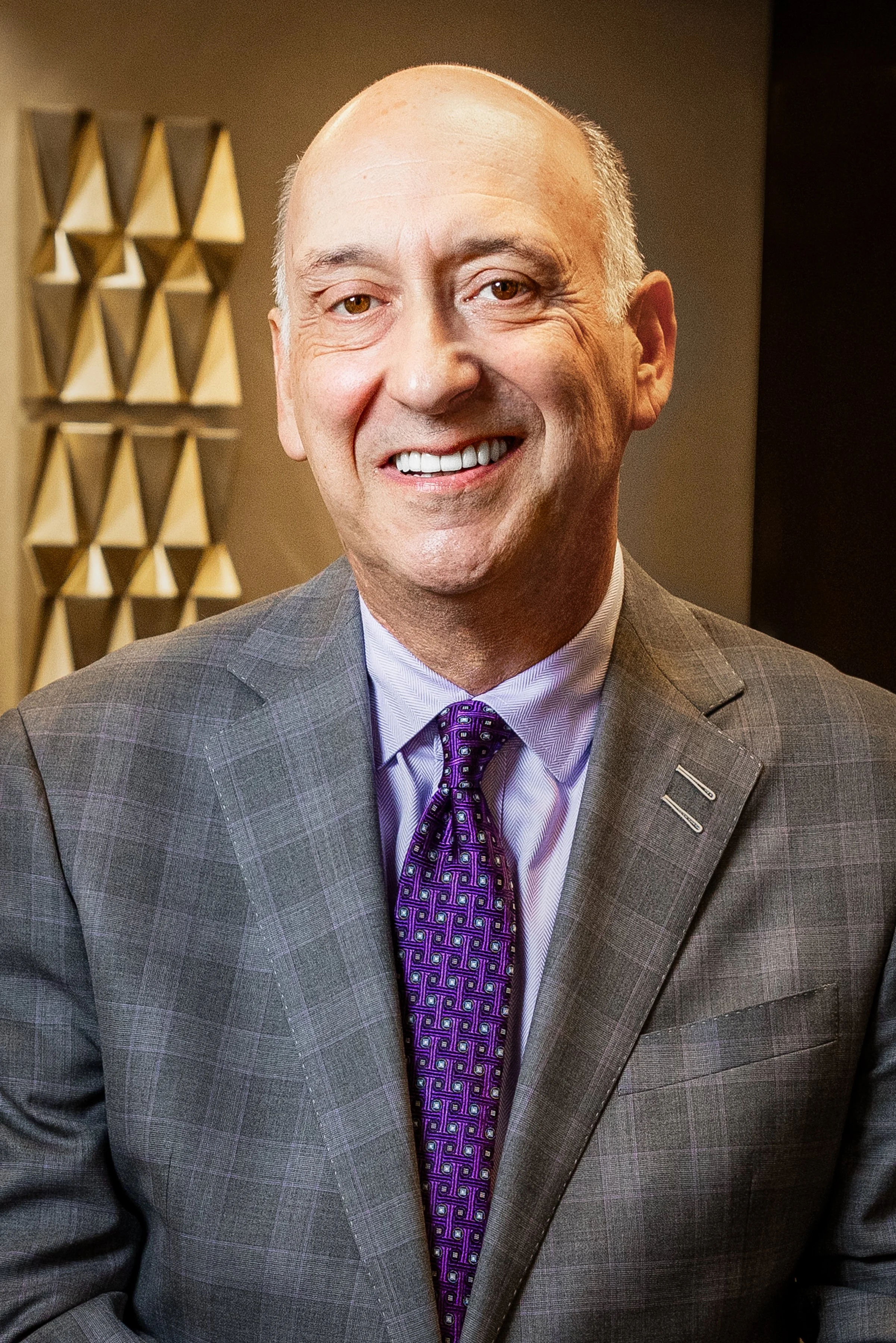 A smiling middle-aged man in a gray suit with a checkered pattern, purple patterned tie, and light purple dress shirt, standing indoors.