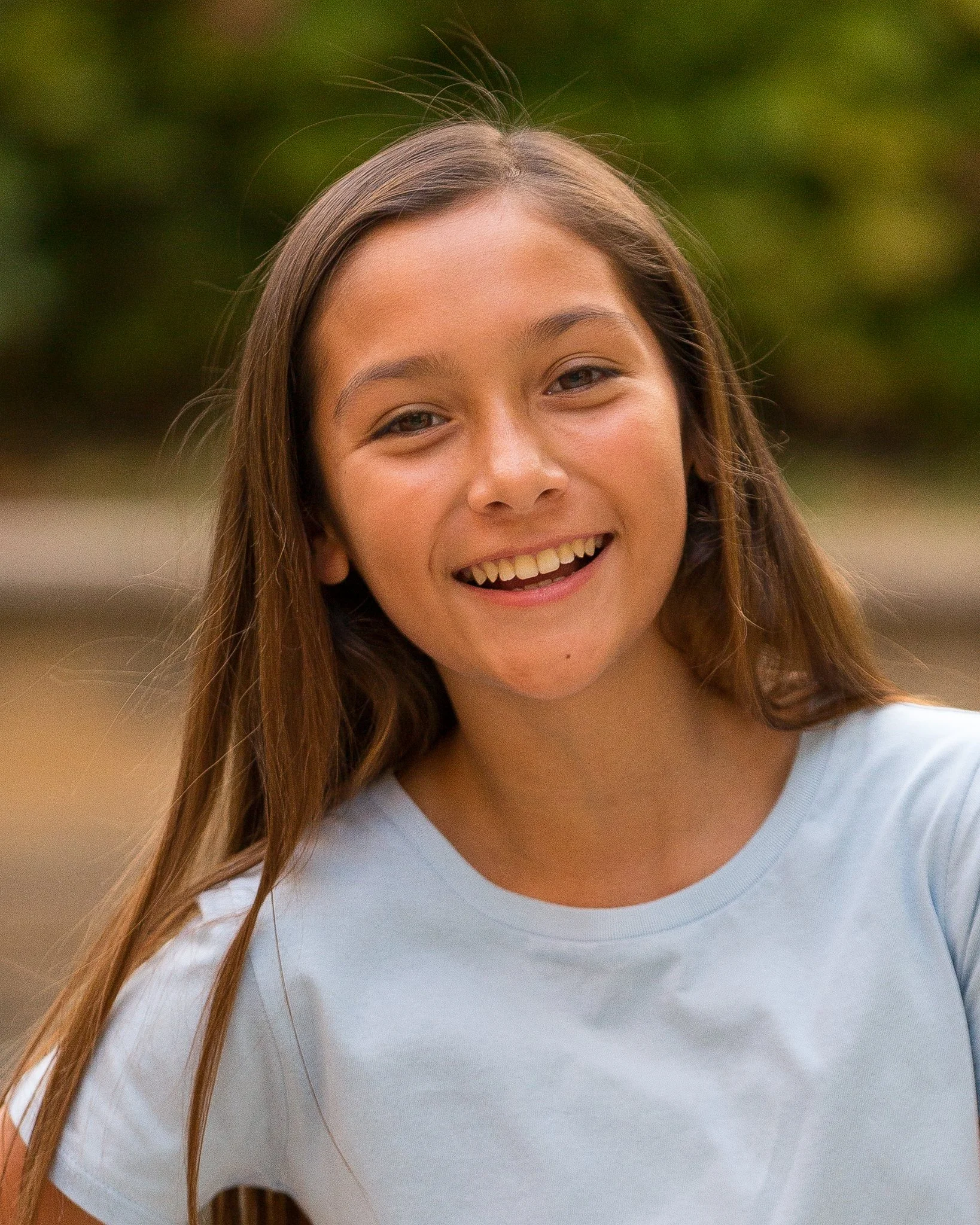 A young girl with long brown hair smiling outdoors, wearing a light blue t-shirt, with blurred greenery in the background.