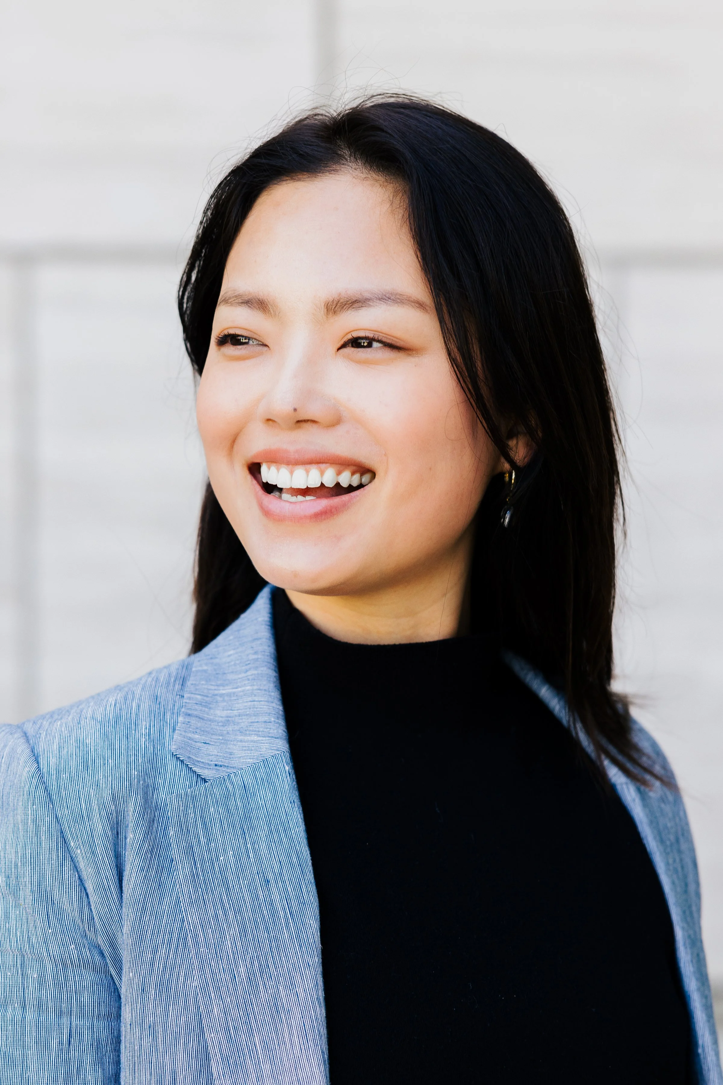 A woman with dark hair smiling in a black top and gray blazer, looking to the side, outdoors.