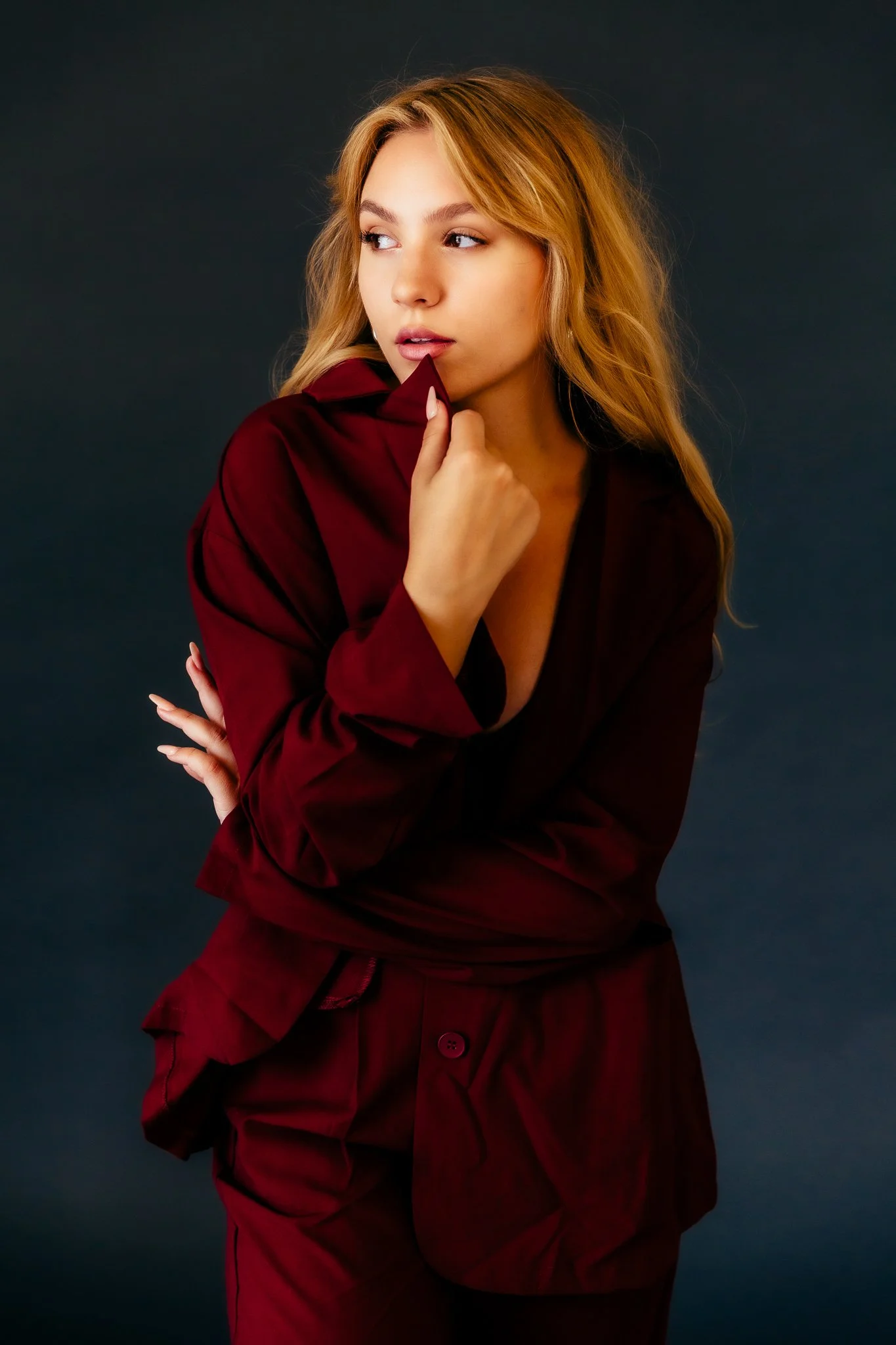 Young woman with blonde hair in a red outfit, standing against a dark background, looking to the side with her left hand near her chin.