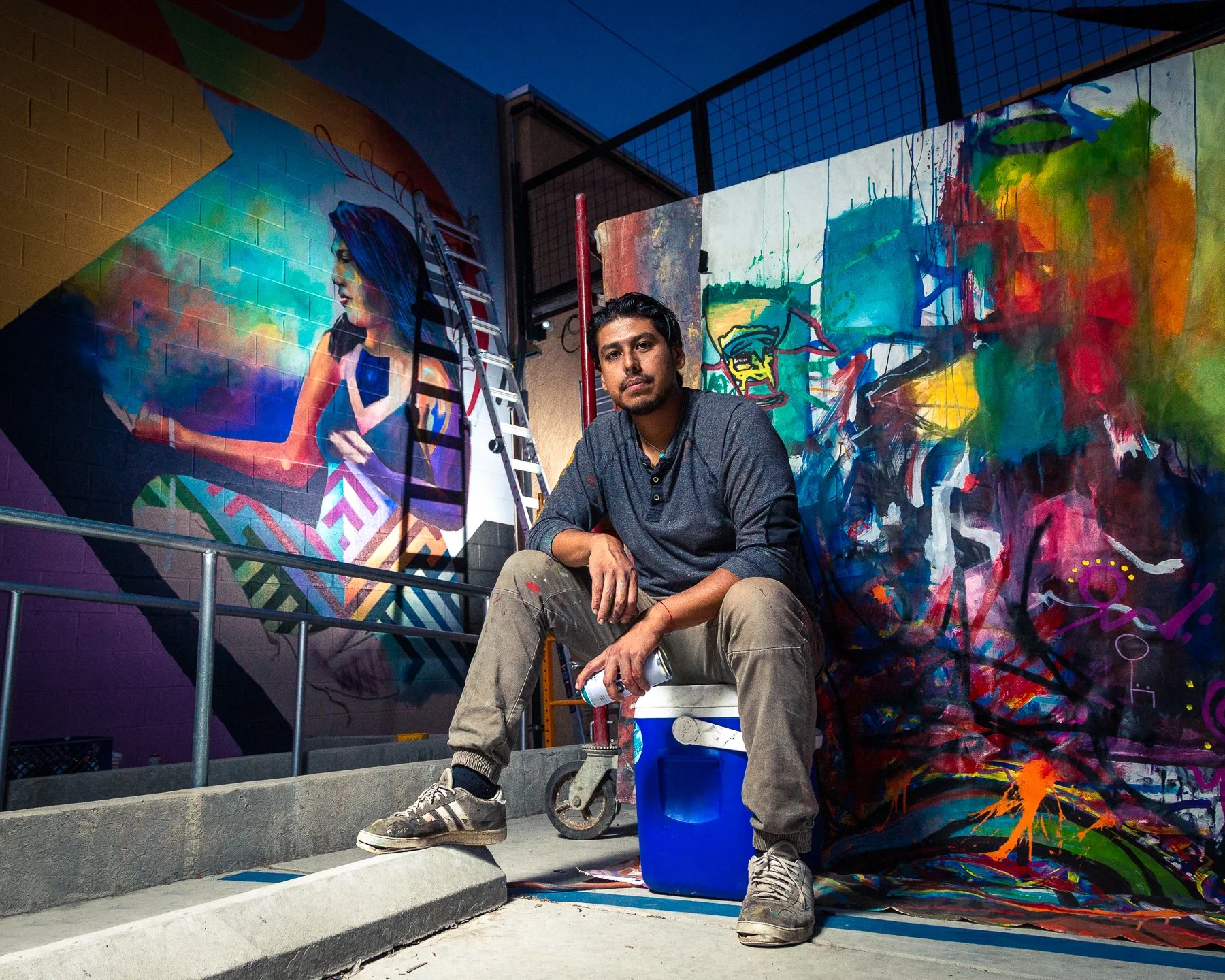 A male artist sitting on a cooler in front of colorful murals, holding a spray paint can, with more murals and a ladder in the background.