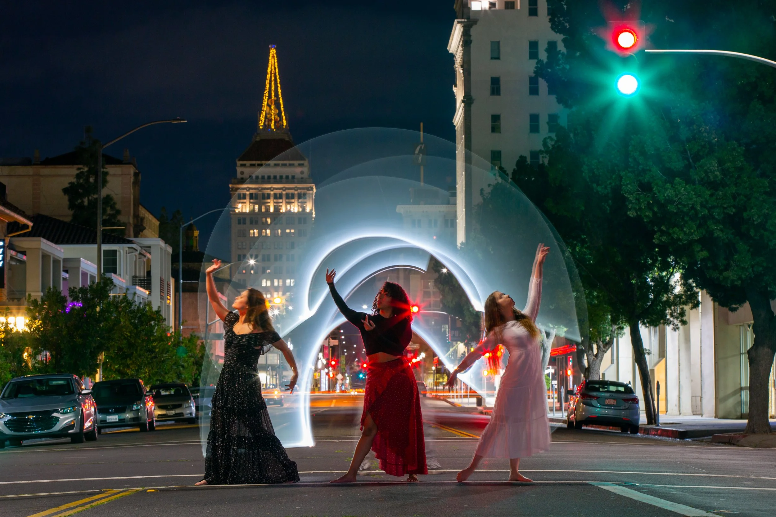 Three women in dresses dancing in the middle of a city street at night, with light trails and a tall building with a pointed, illuminated top in the background.