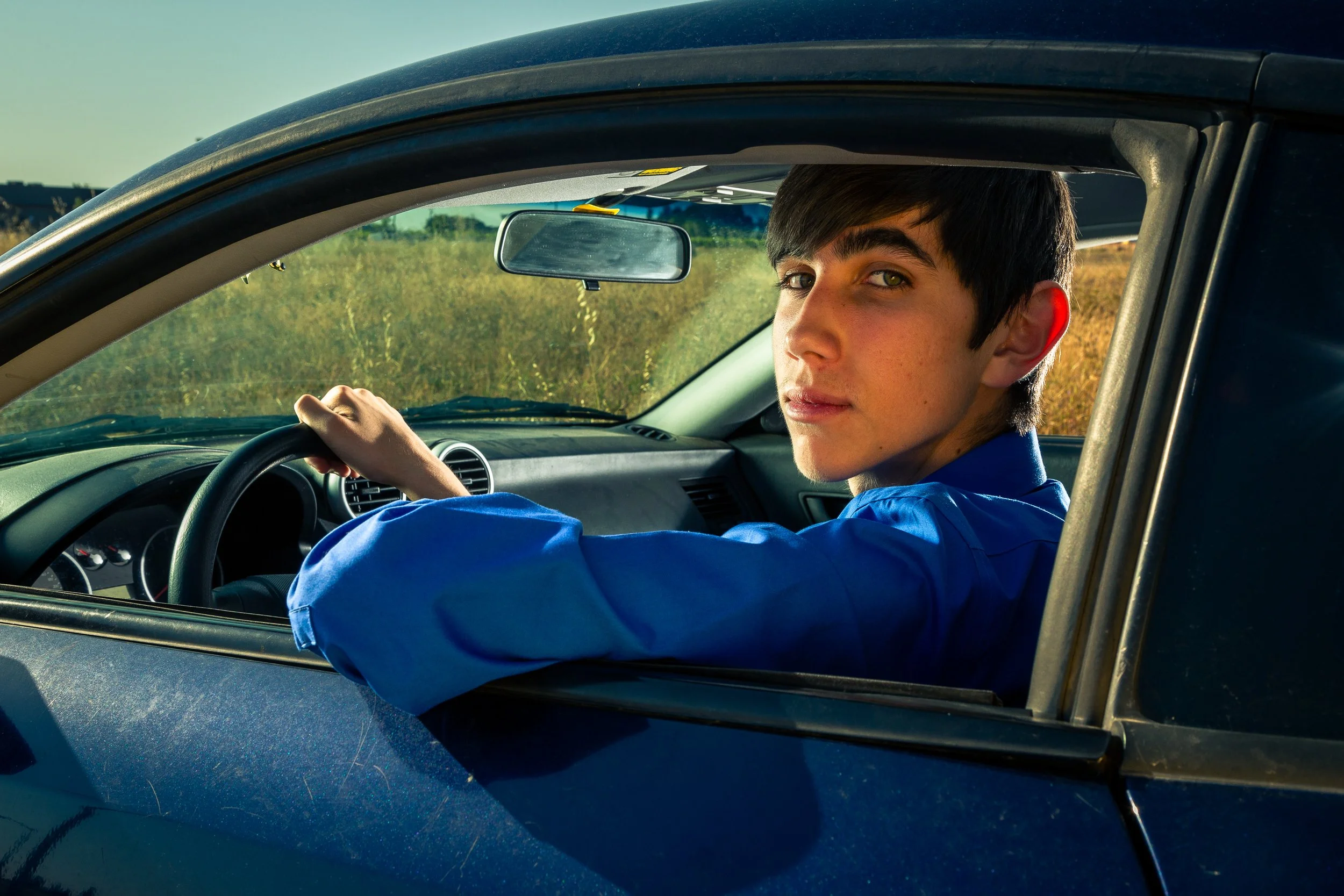 Young man with dark hair wearing a blue shirt sitting in the driver's seat of a car, looking back over his shoulder and holding the steering wheel, with a grassy field and clear sky outside the vehicle.