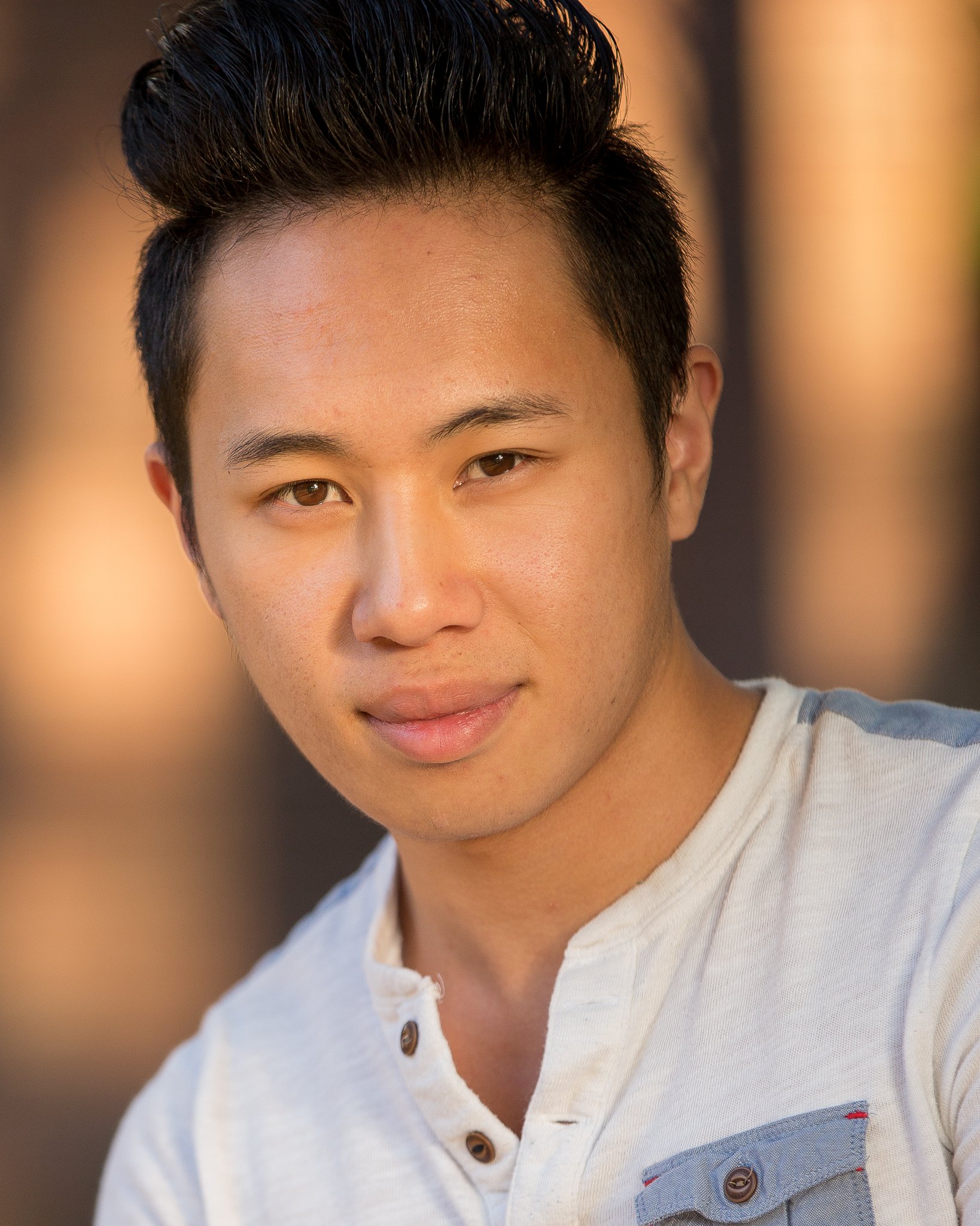 A young man with short black hair and light brown eyes, wearing a white shirt, smiling outdoors at sunset.