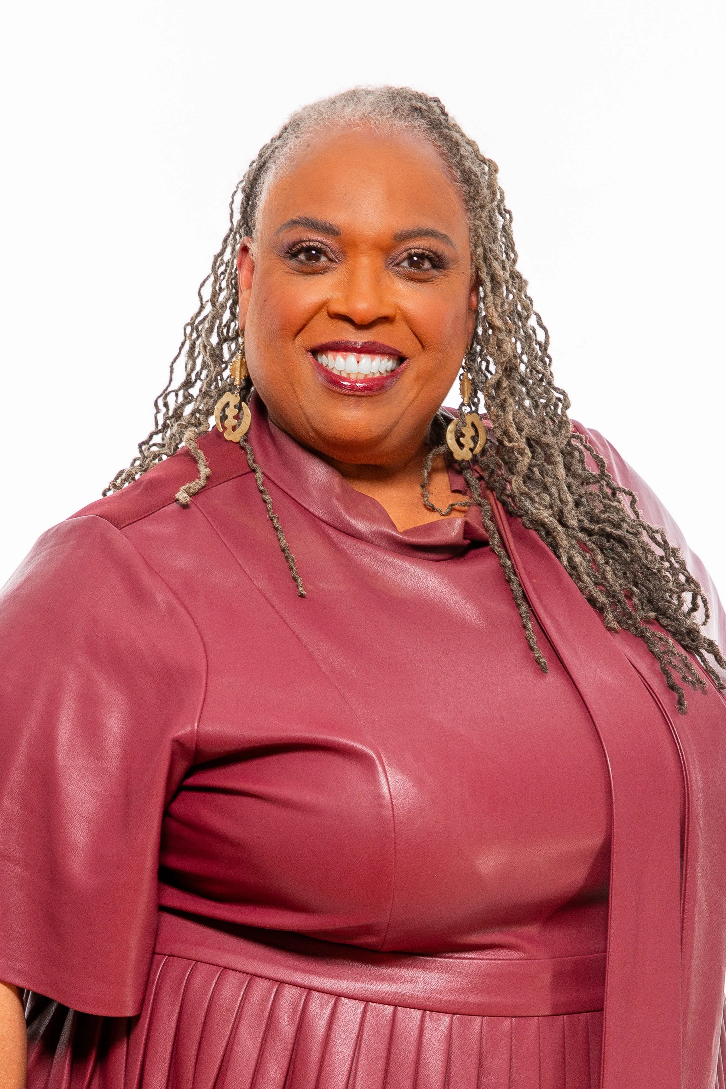 A smiling African American woman with gray dreadlocks wearing a maroon leather dress with zipper detail, gold earrings, and bright lipstick on a plain white background.
