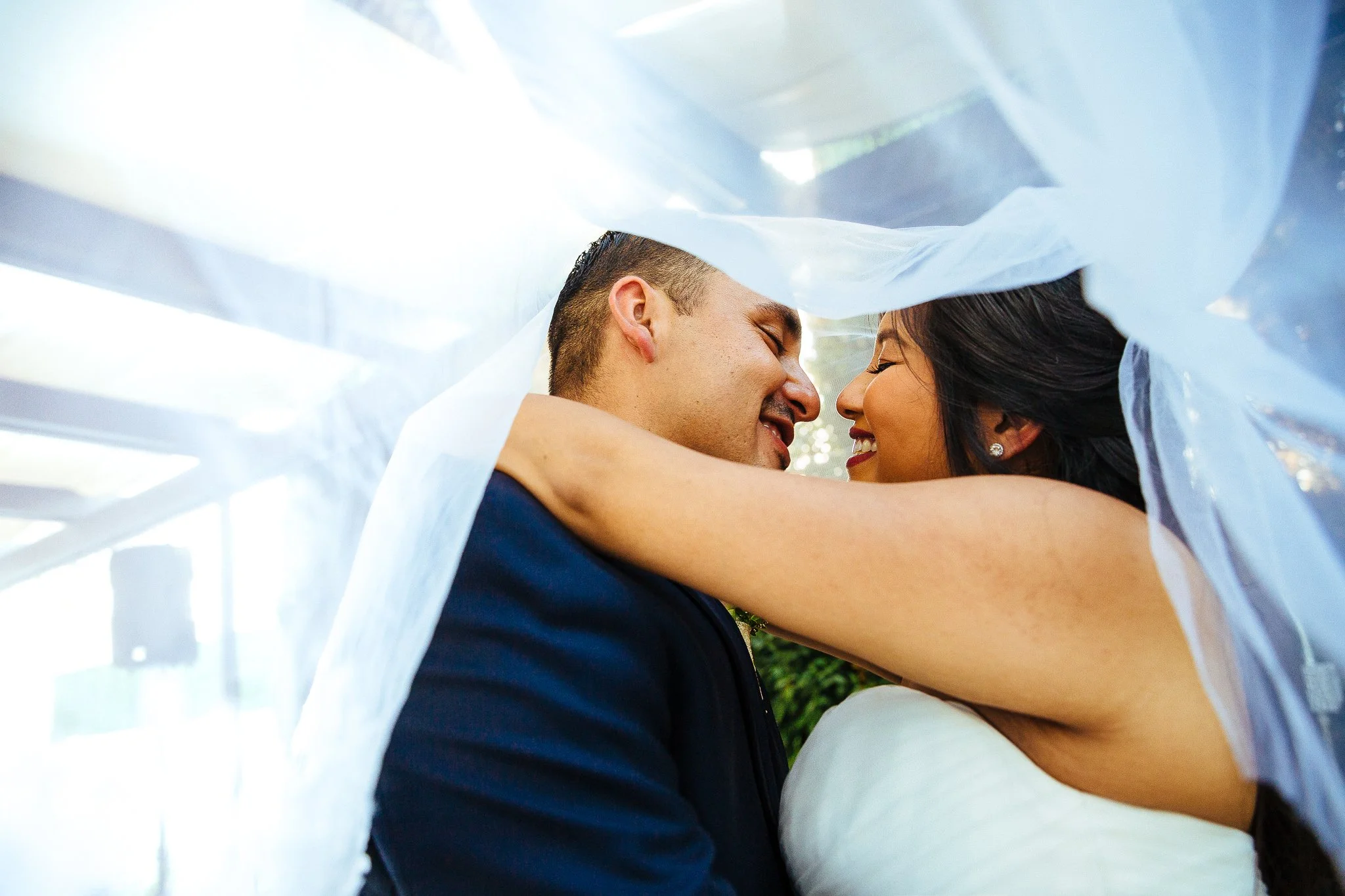 A newlywed couple sharing a close, intimate moment with their foreheads touching, smiling, under a white fabric canopy.