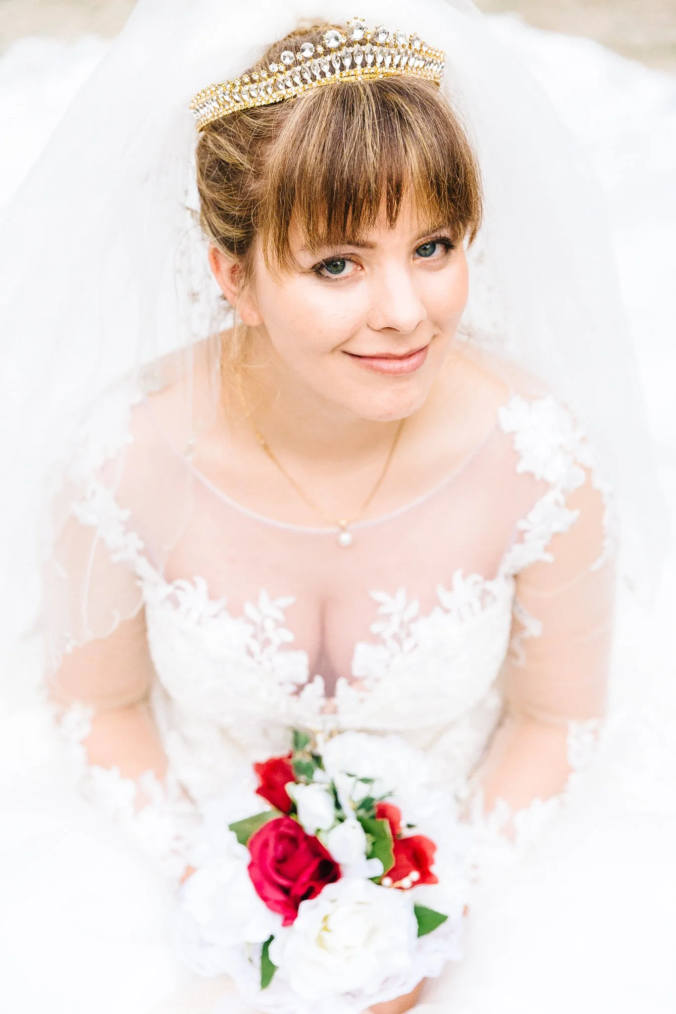 Bride with a pearl and gold crown, wearing a lace wedding dress with floral appliques, holding a bouquet of red and white roses, smiling, with a white veil behind her.