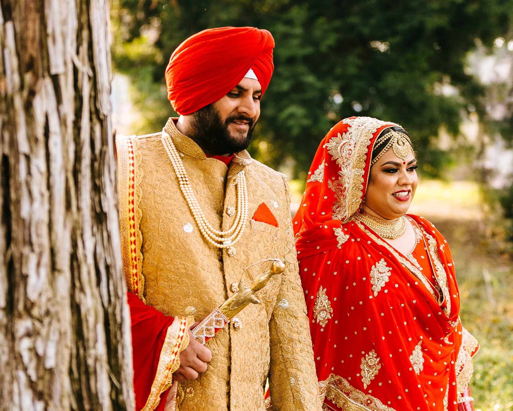 Indian bride and groom in traditional wedding attire standing outdoors behind a tree.