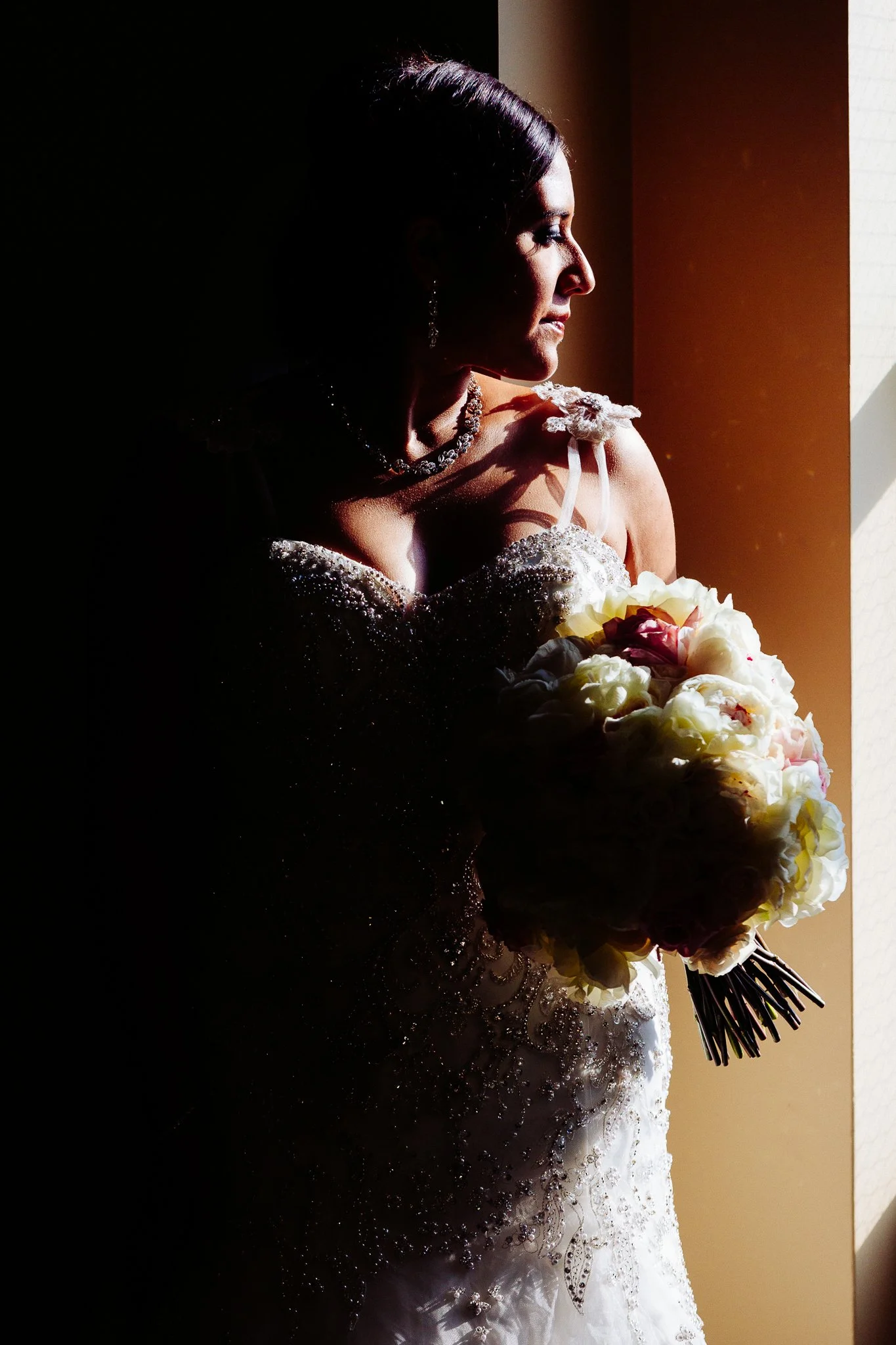 A bride stands by a window, holding a bouquet of white and pink flowers, with sunlight illuminating her face and wedding dress.