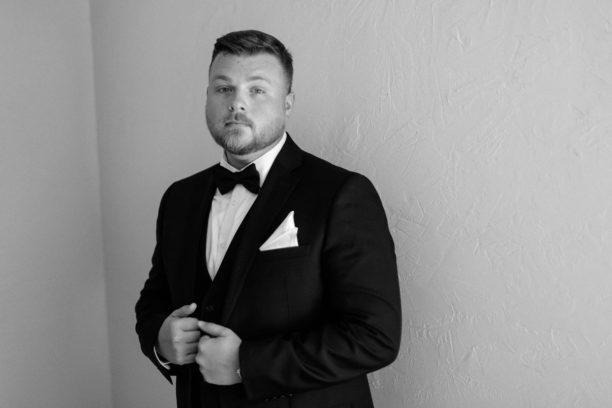 Black and white photo of a man in a tuxedo with a bow tie, white dress shirt, and pocket handkerchief, standing against a textured wall.