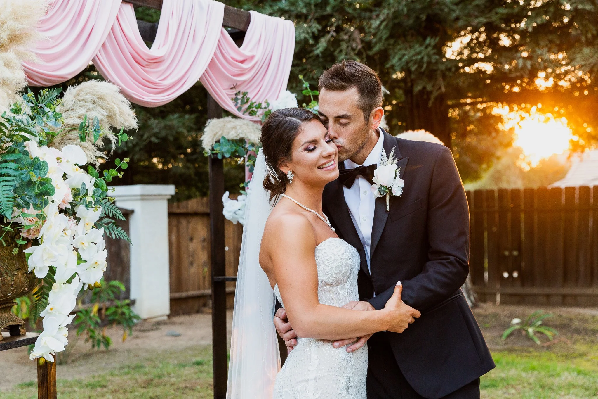 A newlywed couple embracing outdoors at sunset, with the groom kissing the bride on her forehead, wedding decorations, flowers, and pink drapery in the background.