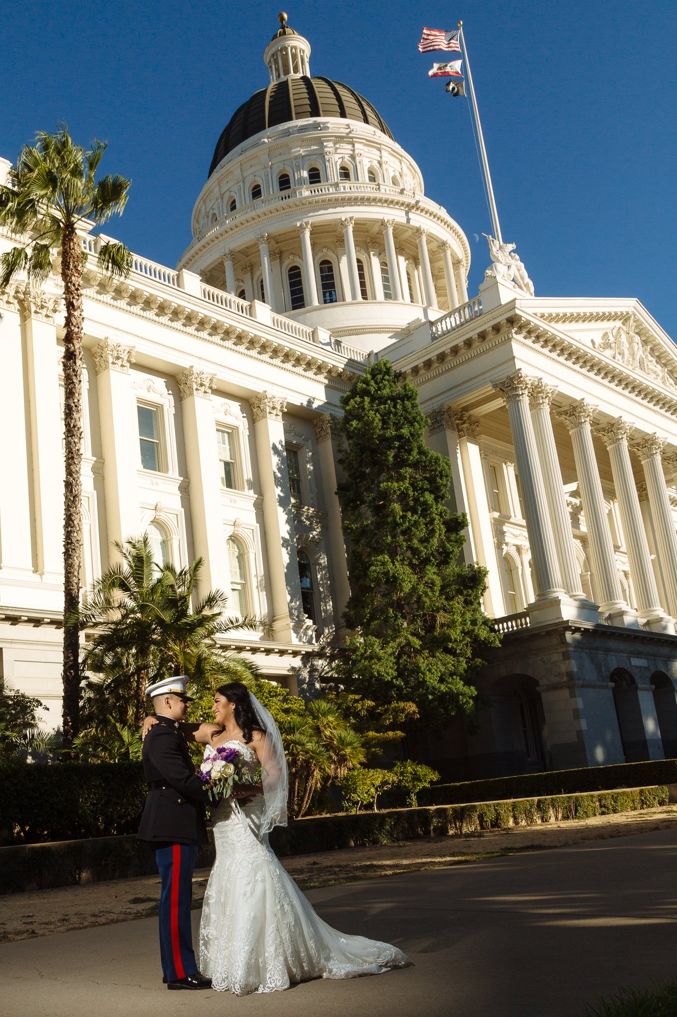 A wedding couple in front of the California State Capitol building with blue sky behind them.