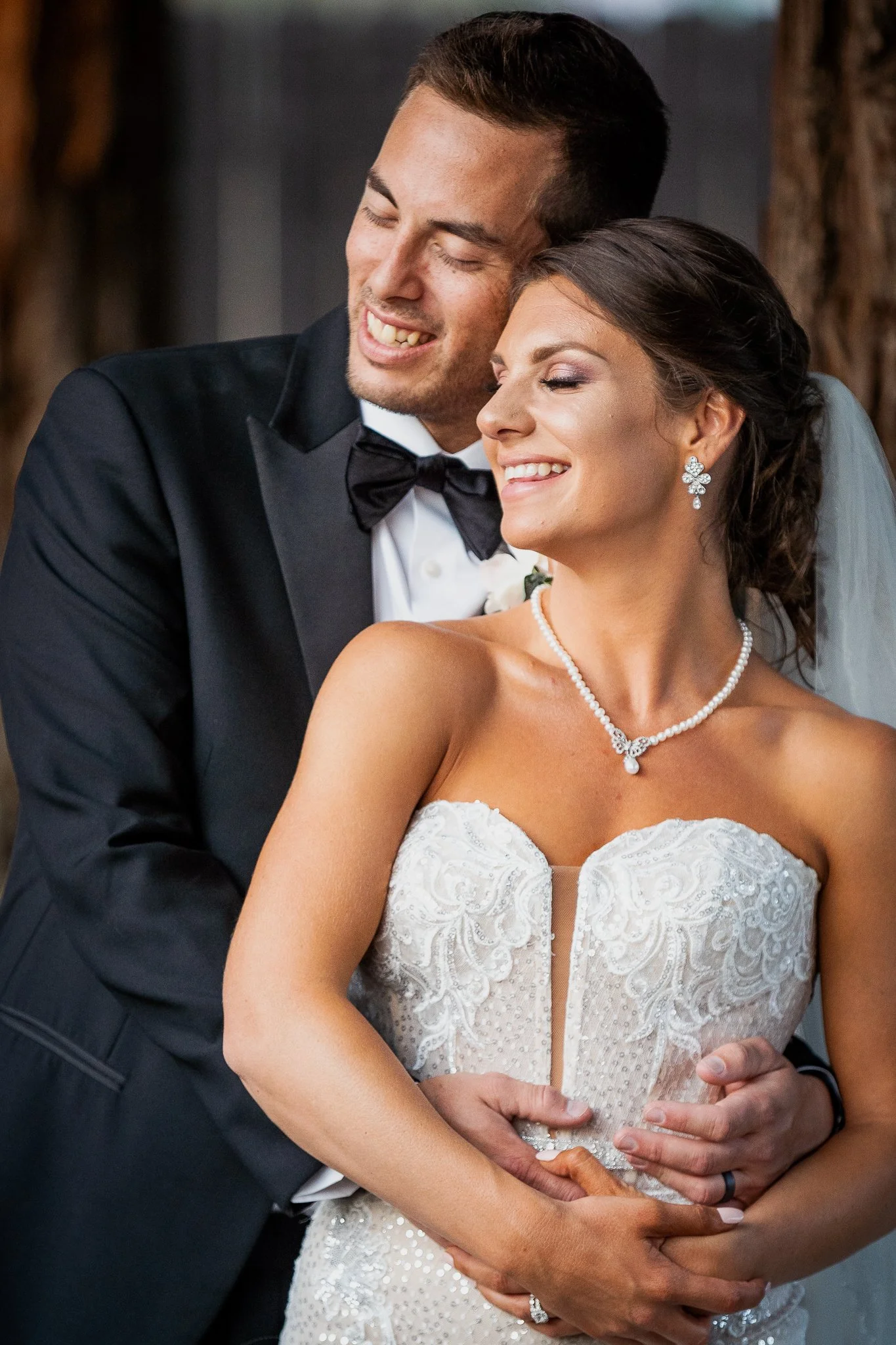 A bride and groom smiling and hugging at their wedding, with the groom in a black tuxedo and the bride in a strapless wedding gown with jewelry.