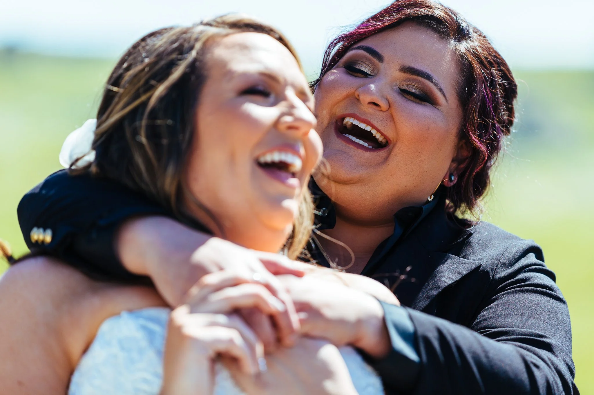 Two women joyfully hugging outdoors on a sunny day, with a blurred green field in the background.