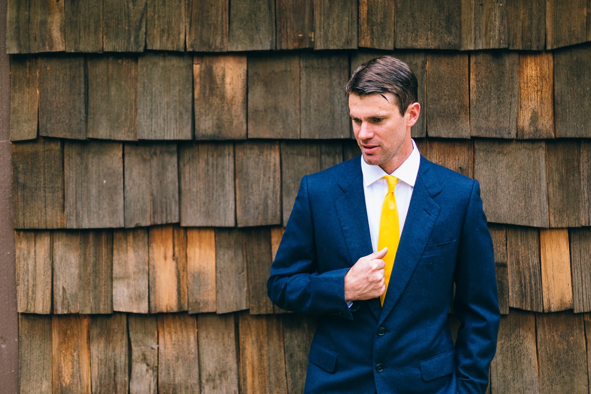 A man in a navy blue suit with a yellow tie standing in front of a wooden shingled wall, looking down thoughtfully.