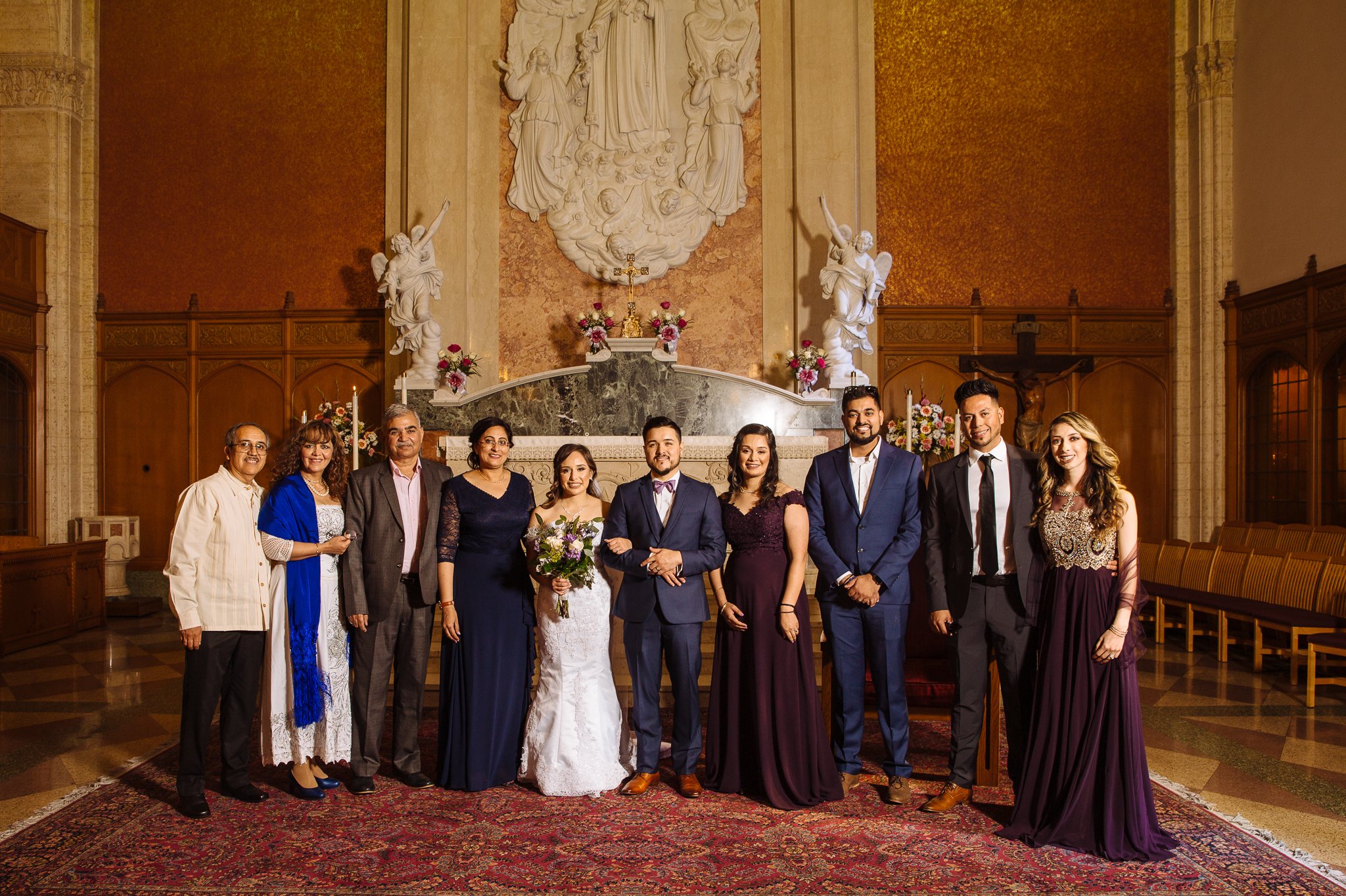 A group of eleven people dressed in formal attire standing in a church with religious statues and artwork, for a wedding celebration.