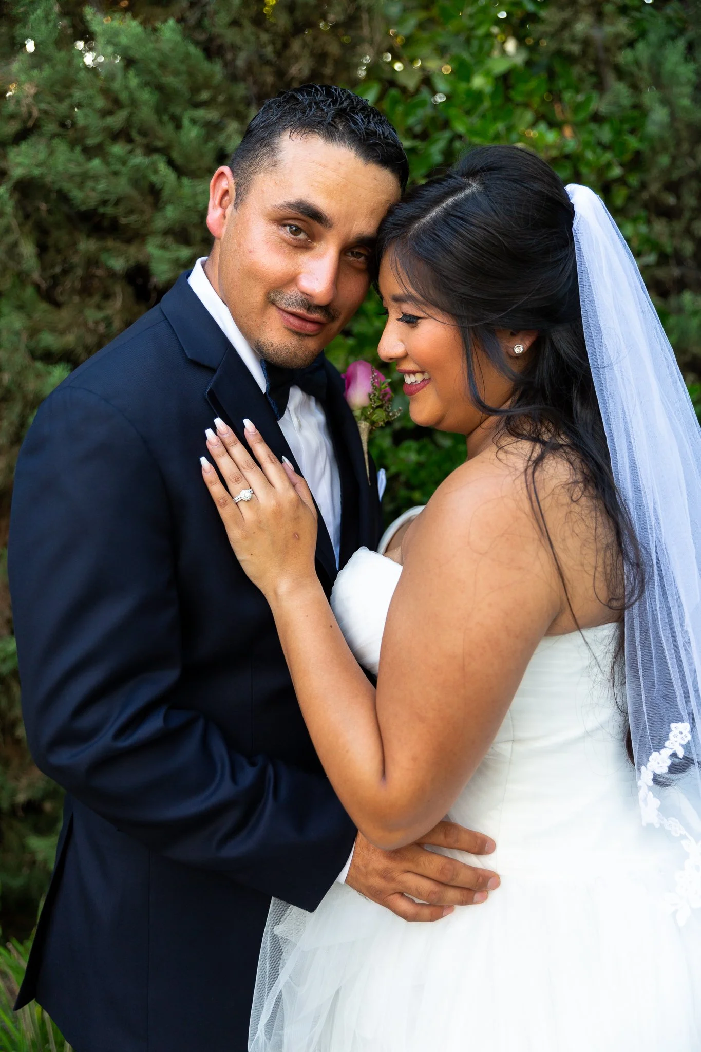 A newlywed couple, dressed in wedding attire, standing close together outdoors with greenery in the background. The bride is smiling, showing her ring, while the groom has a slight smile.
