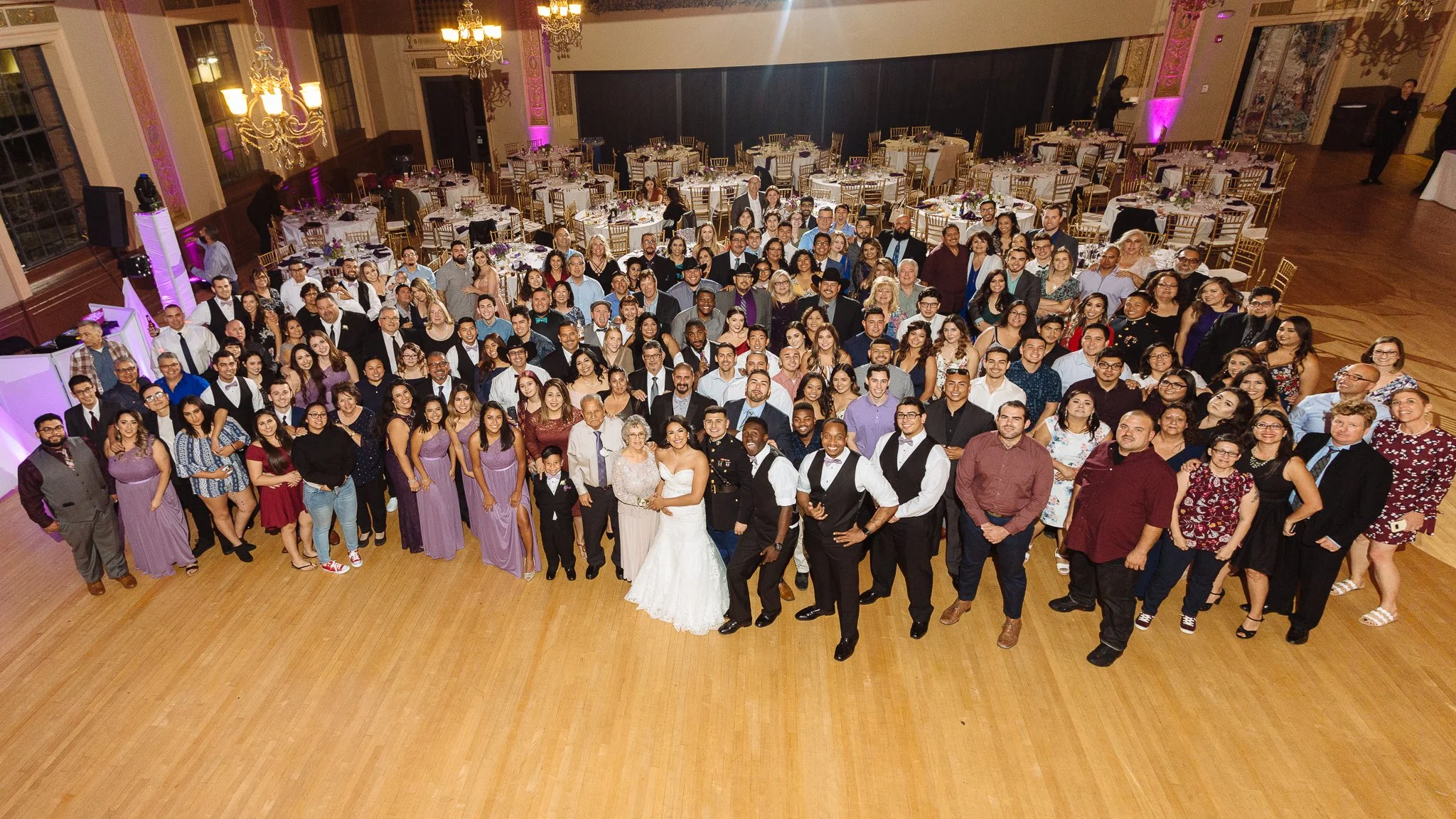 Group of people gathered for a celebration in a grand ballroom with decorated tables and chandeliers, with a bride and groom at the center.