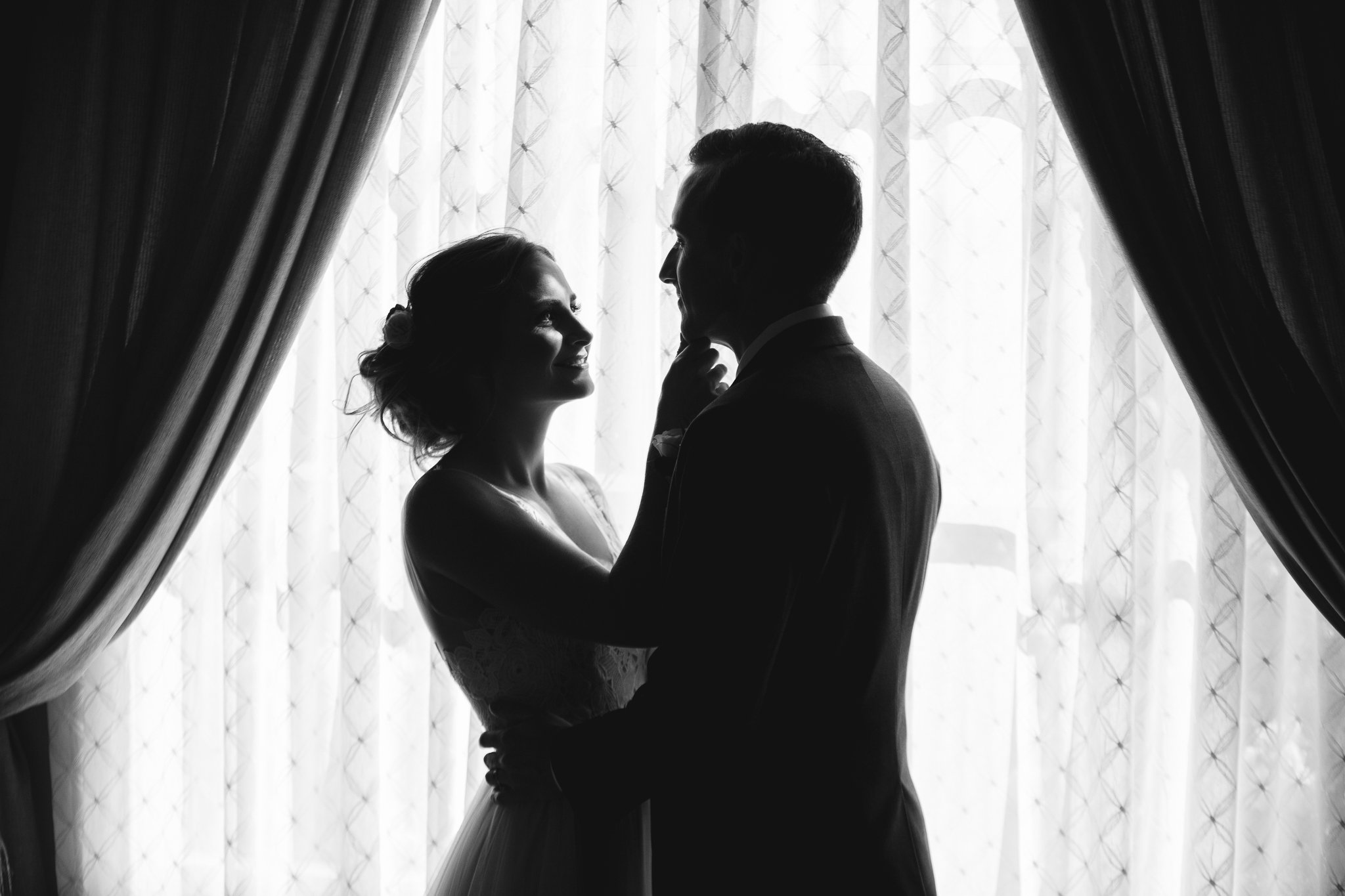 A black and white photo of a bride and groom standing close together in front of a backlit curtain, with the bride smiling at the groom as he gently touches her chin.