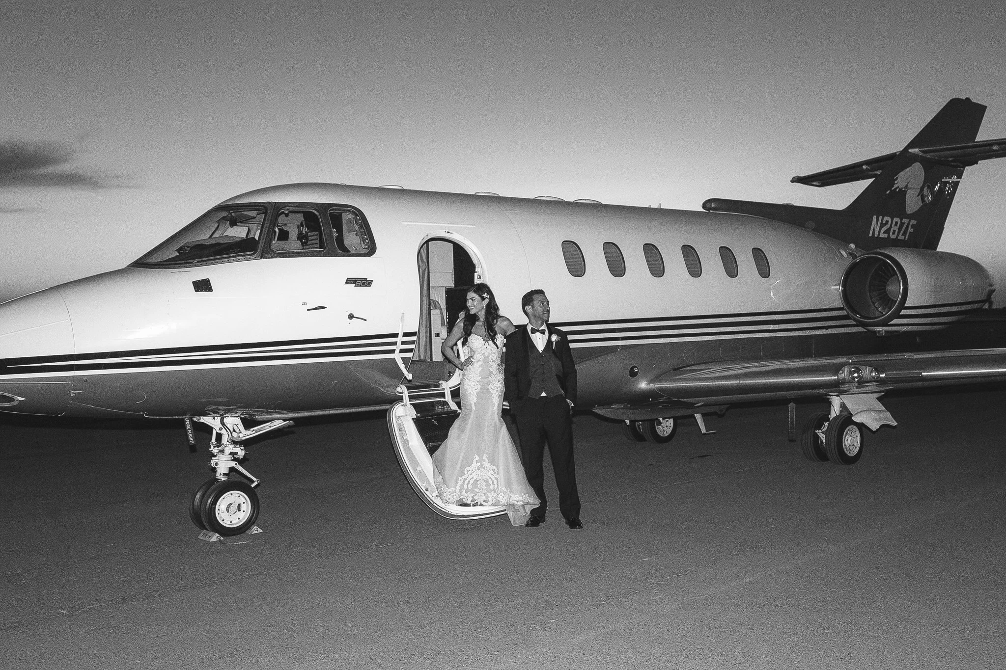 Black and white photo of a bride and groom standing by a private jet on an airport tarmac during sunset or dusk.