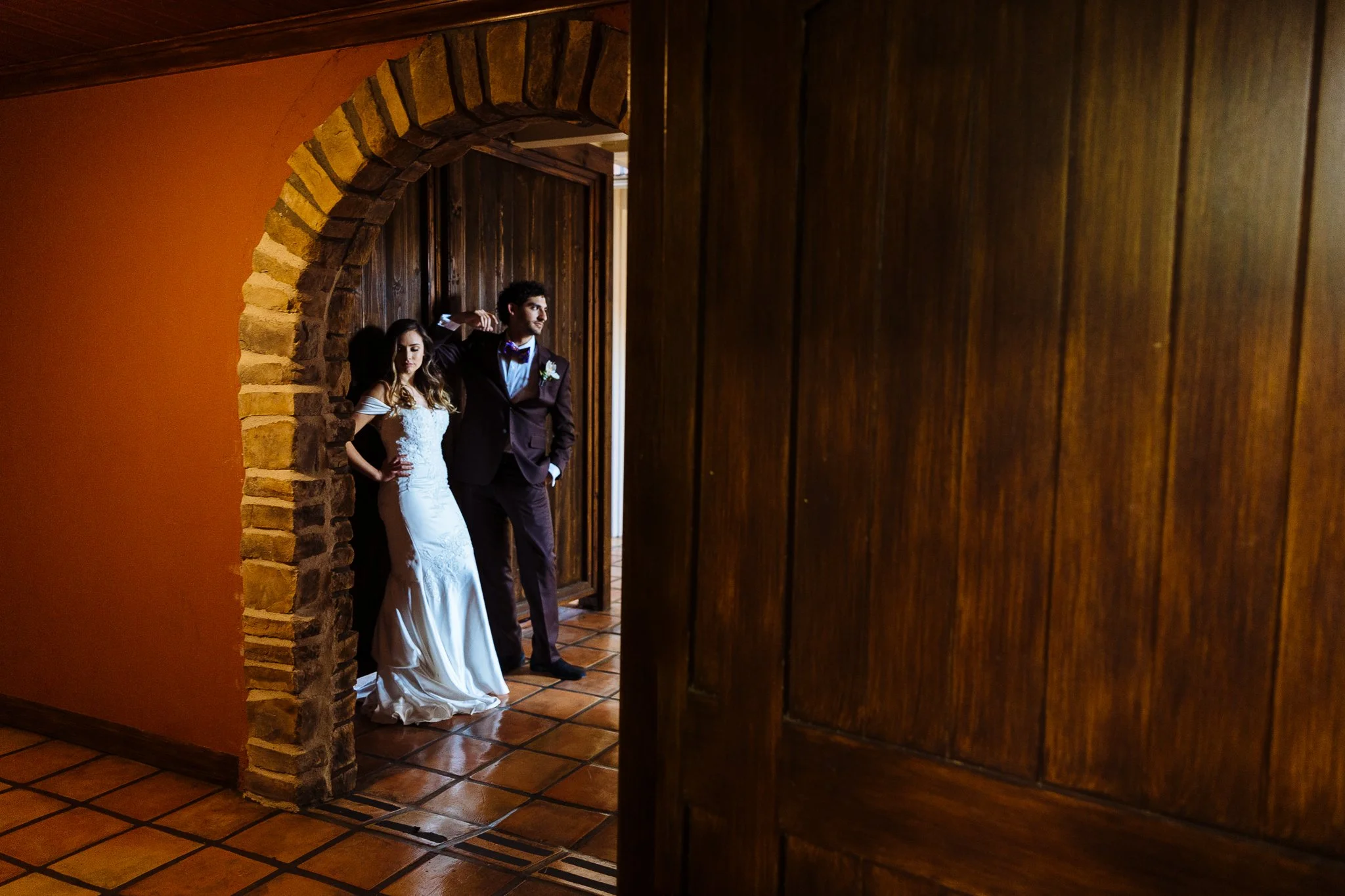 A bride and groom standing in a doorway inside a warmly lit room with wooden walls and tiled floor, the bride in a white wedding gown and the groom in a dark suit, both posing for a photo.