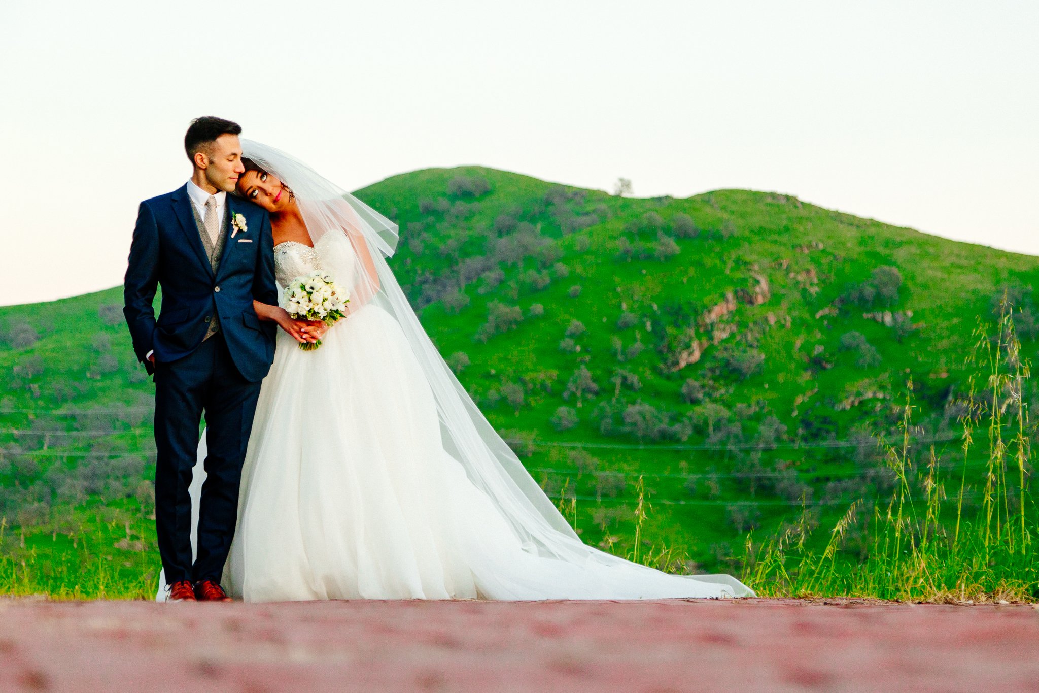 A bride and groom standing close together outdoors with green hills in the background on a clear day.