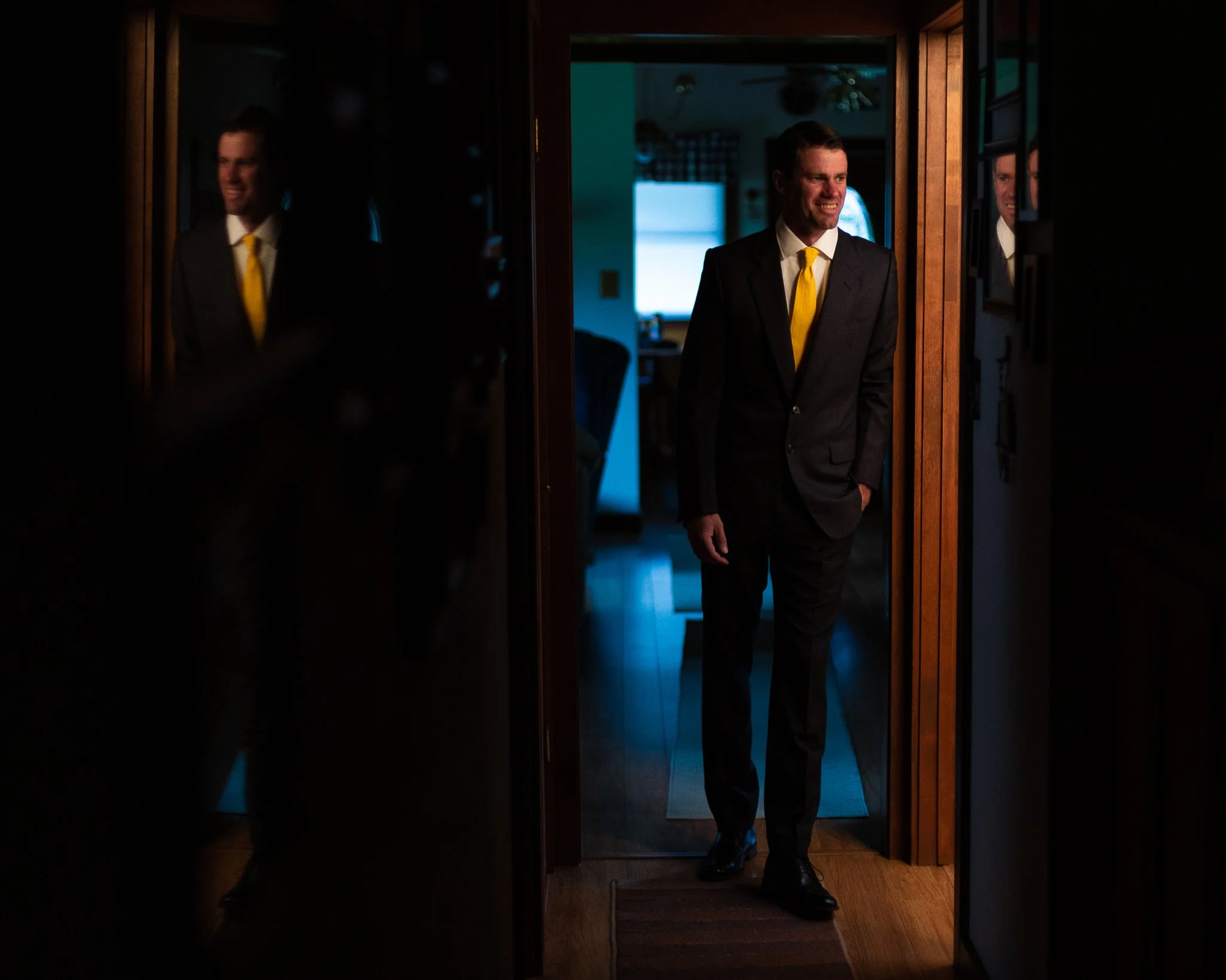 A man in a black suit with a yellow tie standing in a doorway, smiling, with his reflection visible in the mirror on the right and a dimly lit room in the background.