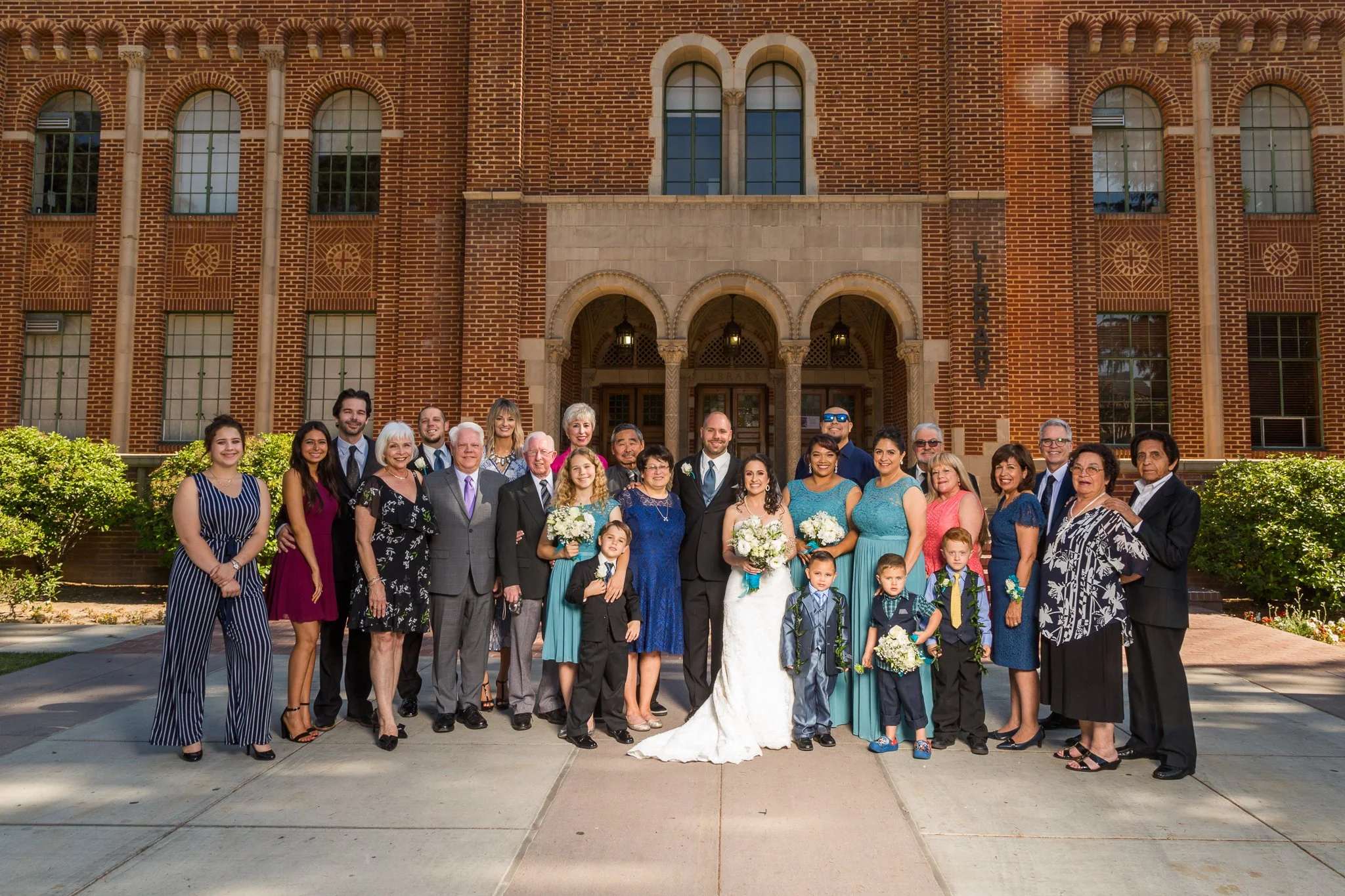 A group of people posing in front of a red brick building on a sunny day, likely a wedding celebration.