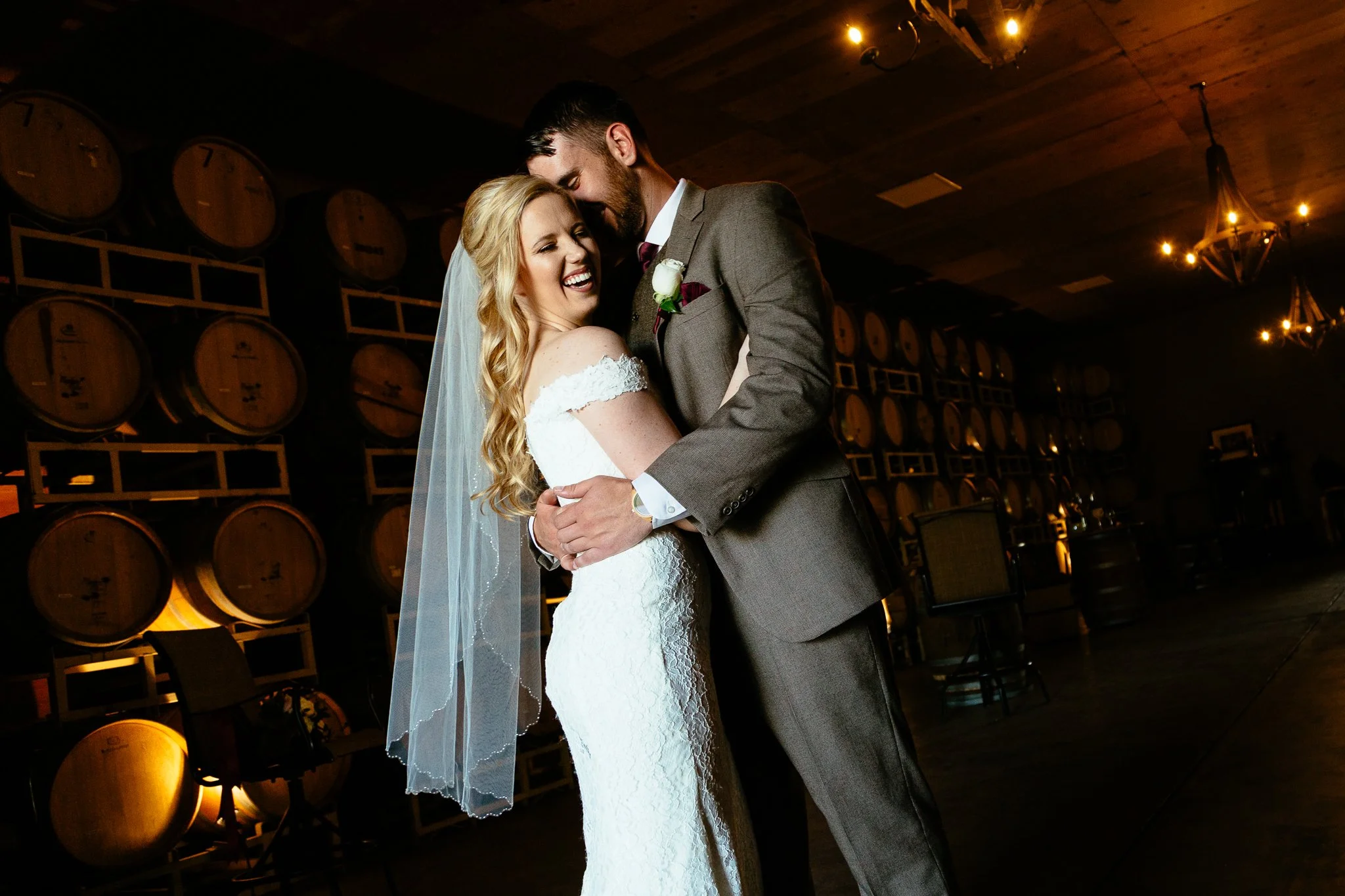 A bride and groom dancing and smiling at their wedding reception, with wine barrels in the background.
